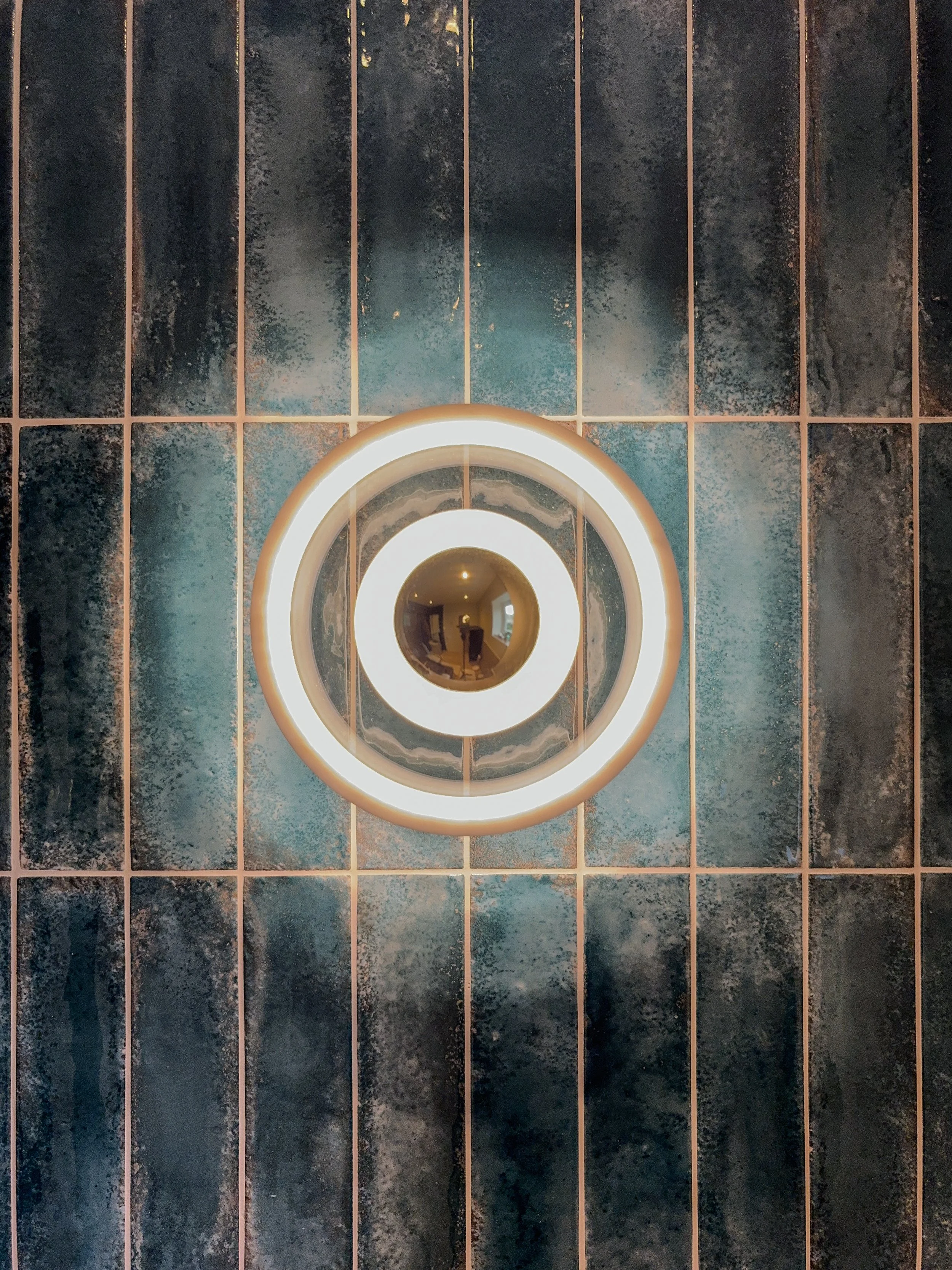 Circular mirror with illuminated rings mounted on a textured dark wall with vertical grid lines.