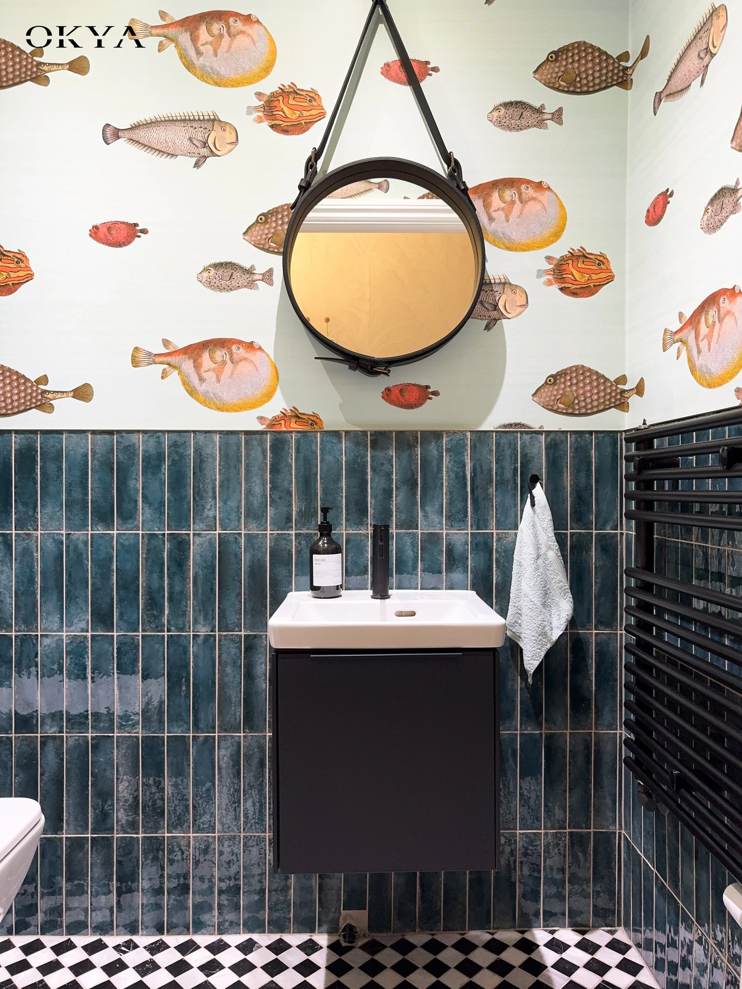 This is where the project takes its biggest risk and earns its biggest reward. Illustrated fish wallpaper on a pale ground, deep teal zellige tiles laid vertically, a leather-strap mirror, black vanity and monochrome marble floor. On paper it should 