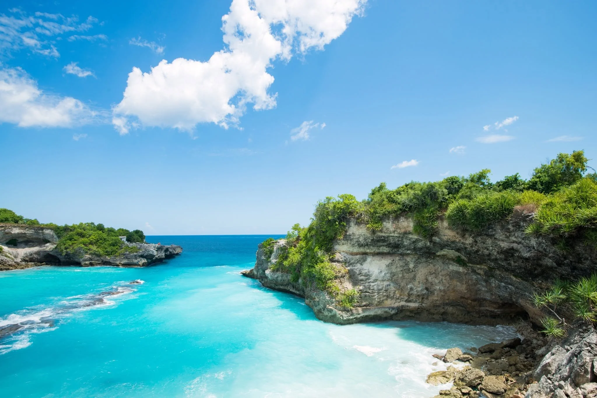 Aerial view of a coastal area with several houses, surrounded by clear turquoise waters and green vegetation. A wooden deck extends over the water, with a small gazebo and chairs. The landscape includes rocky shores and sandy patches.