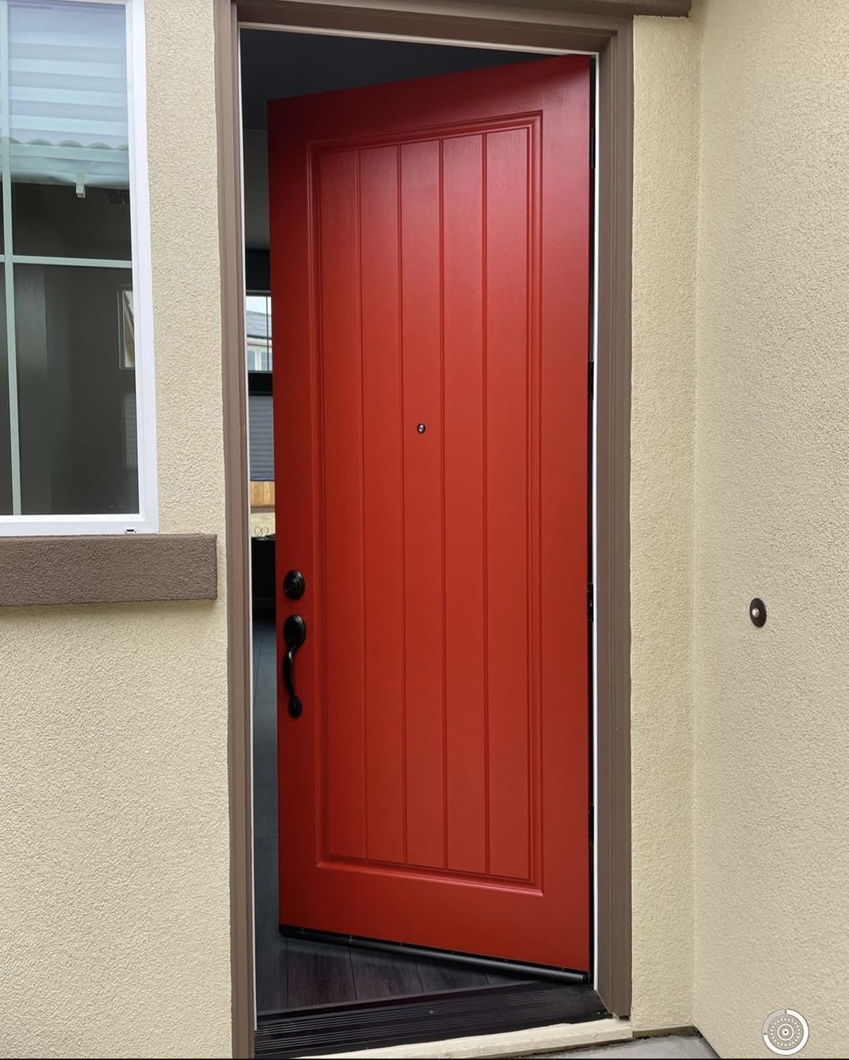 Open red front door with black handle and lock, beige house wall with a window on the left, and a small round doorbell on the right.