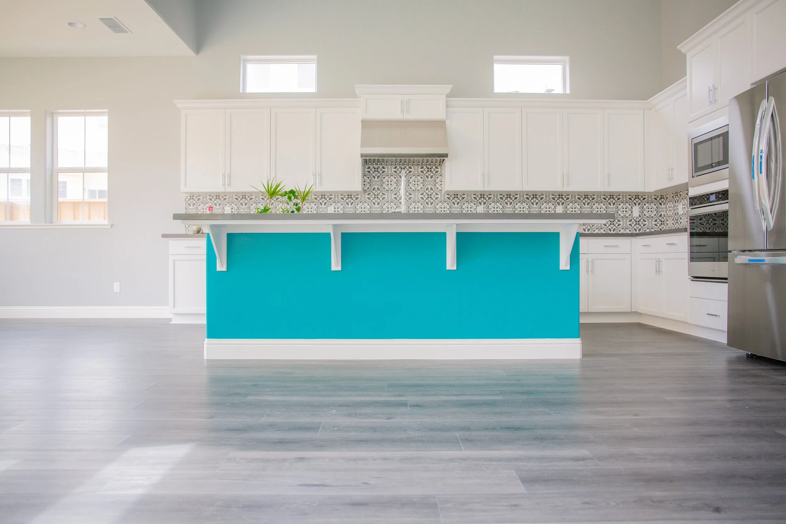 Modern kitchen with white cabinets, patterned tile backsplash, blue kitchen island, stainless steel appliances, gray flooring, and three windows letting in natural light.