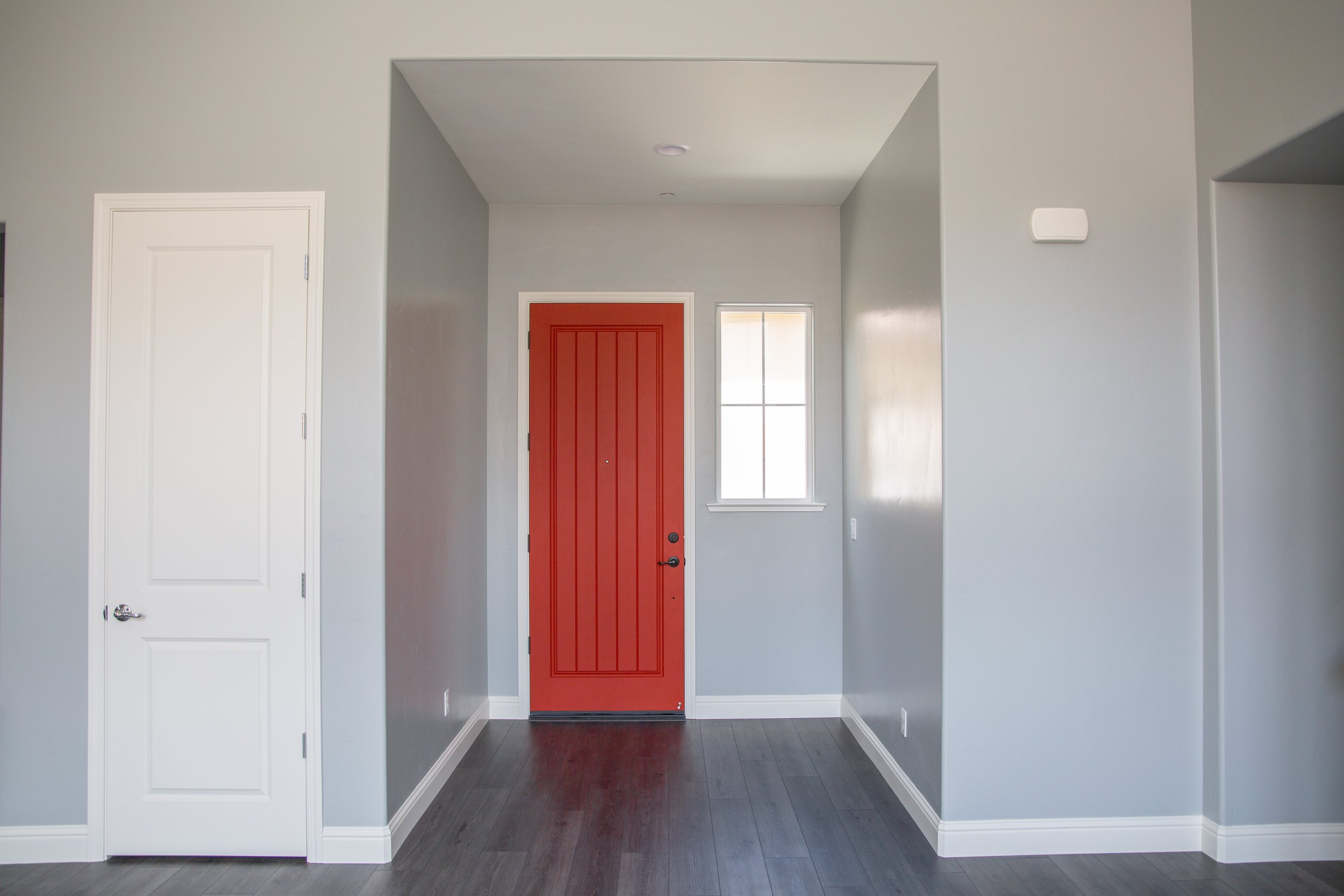 Interior of a modern house entryway with a bright red front door, a white door on the left, and a small window on the right, with gray walls and dark wood flooring.