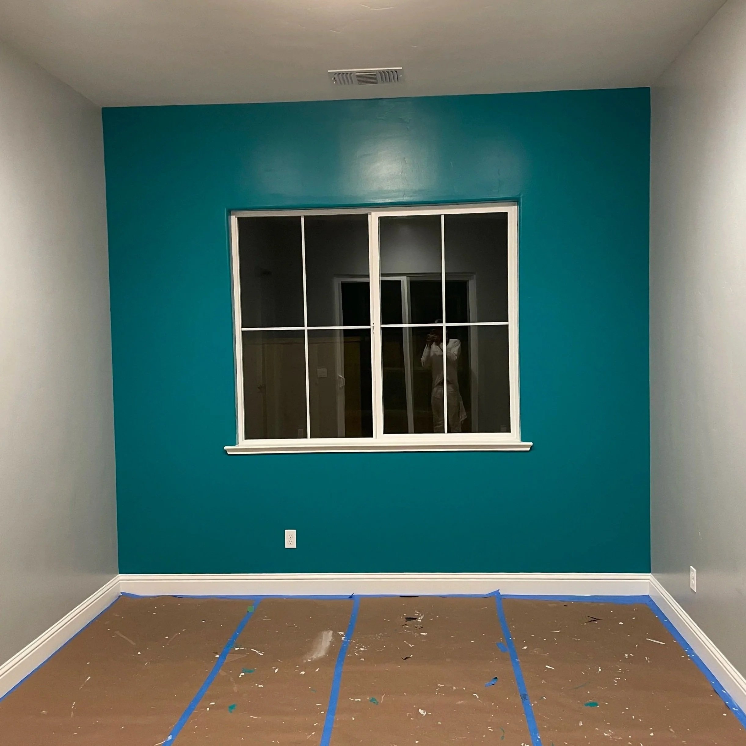 Empty room with blue painted wall, window, and brown floor covered with painter's tape and paint splatters.