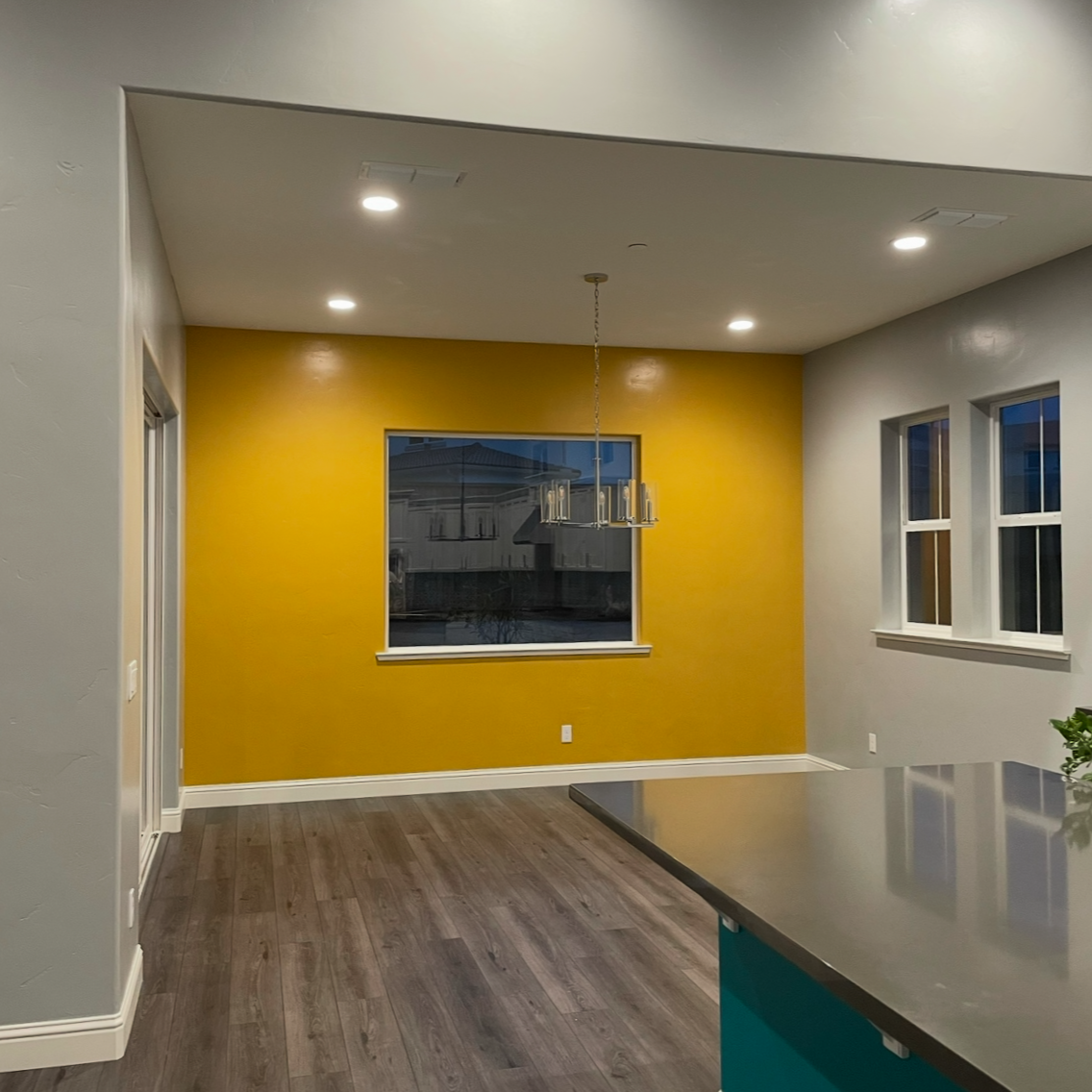 Interior view of a modern dining area with a yellow accent wall, two windows, a hanging chandelier, wood flooring, and a partial view of a kitchen island with a silver countertop