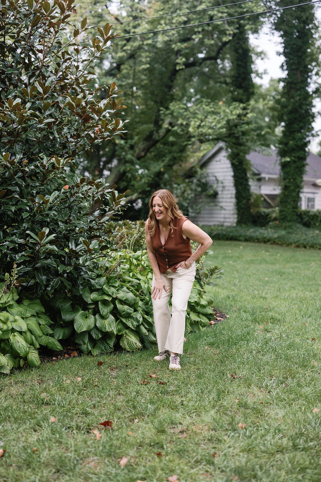 A woman with red hair, wearing a sleeveless brown top, cream-colored pants, and sneakers, laughing while walking through a garden with lush green plants and trees.