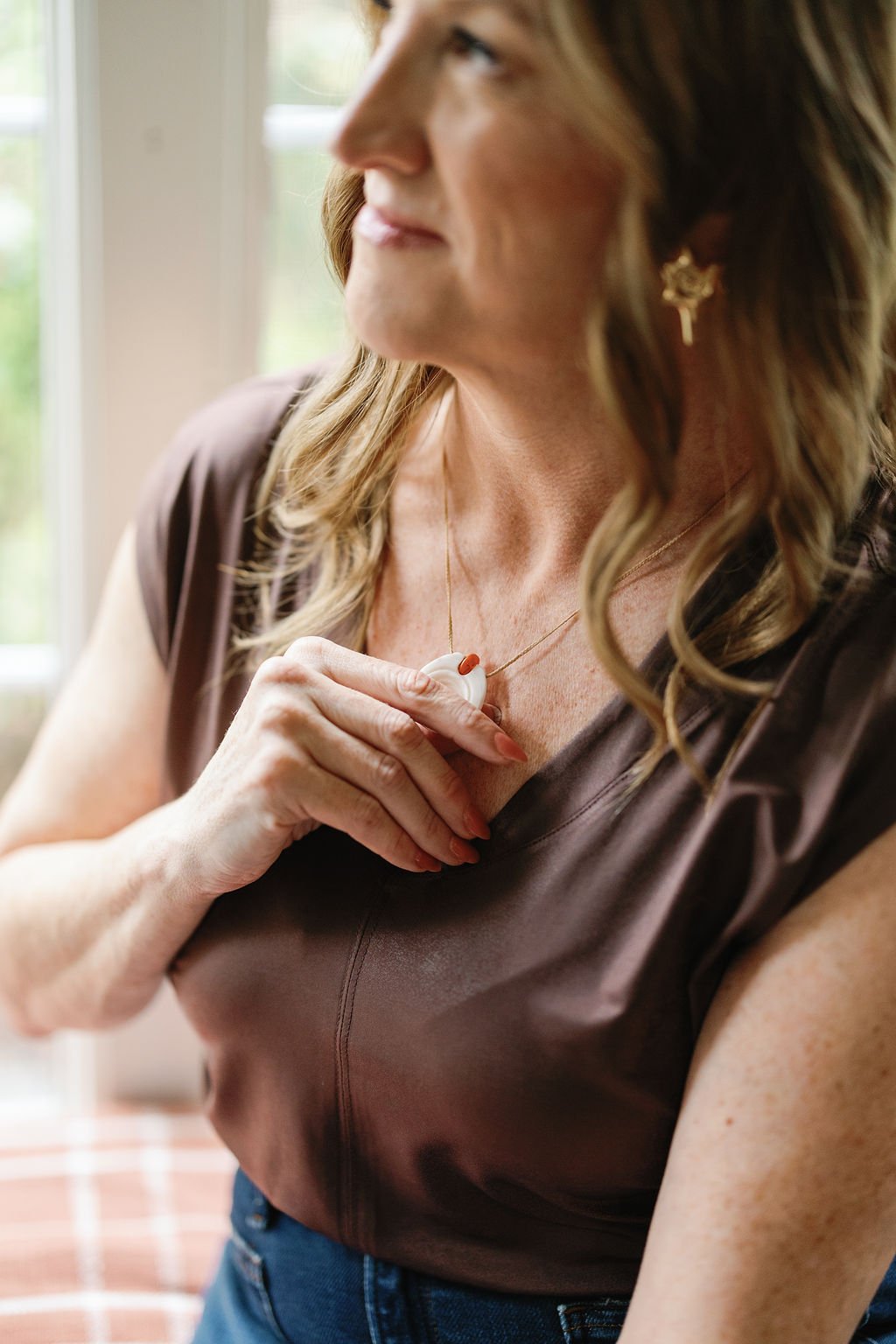 A woman with blonde hair and gold earrings is touching a necklace with a white stone pendant.