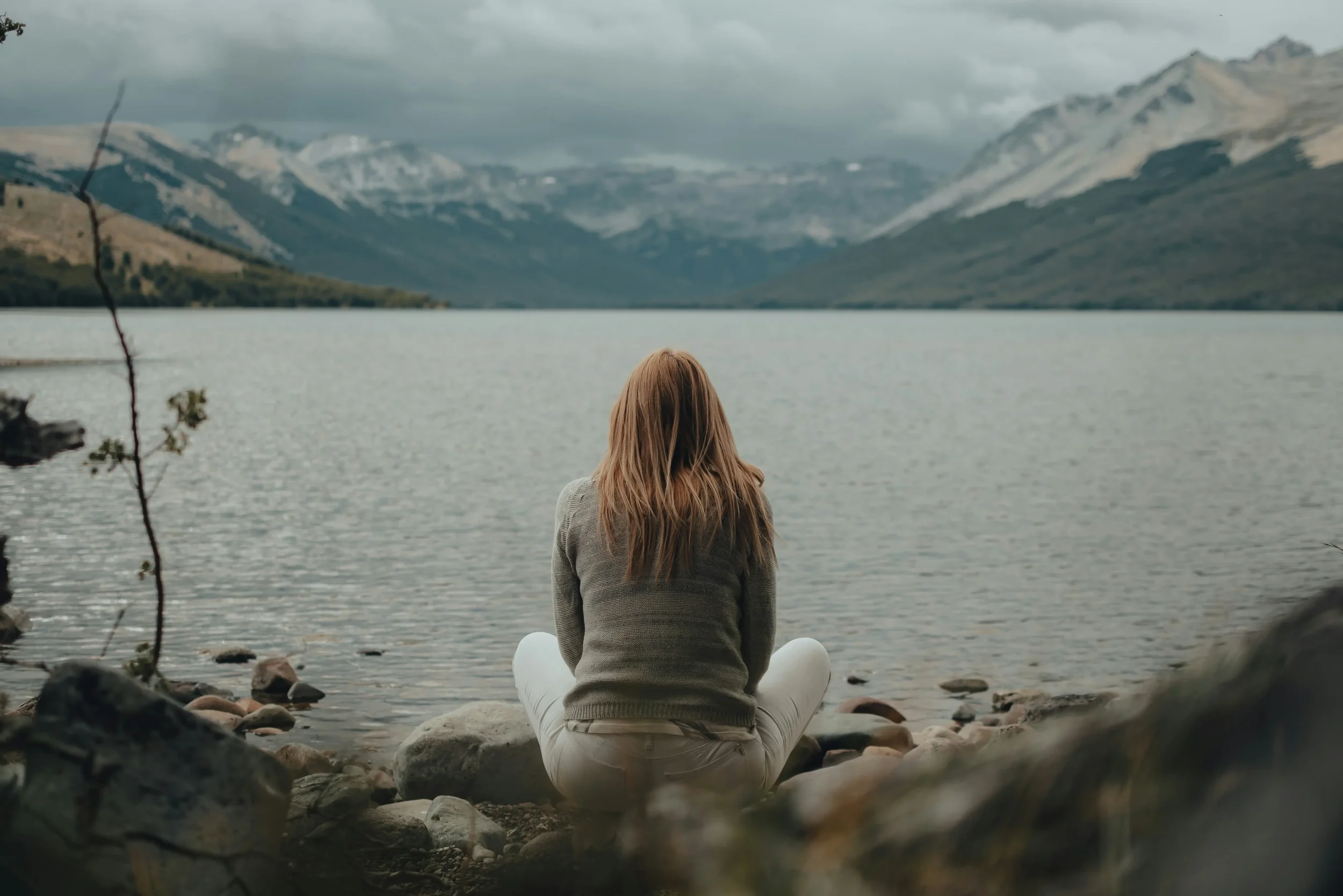 A woman sitting cross-legged on rocks by a lake, overlooking mountains under cloudy sky.