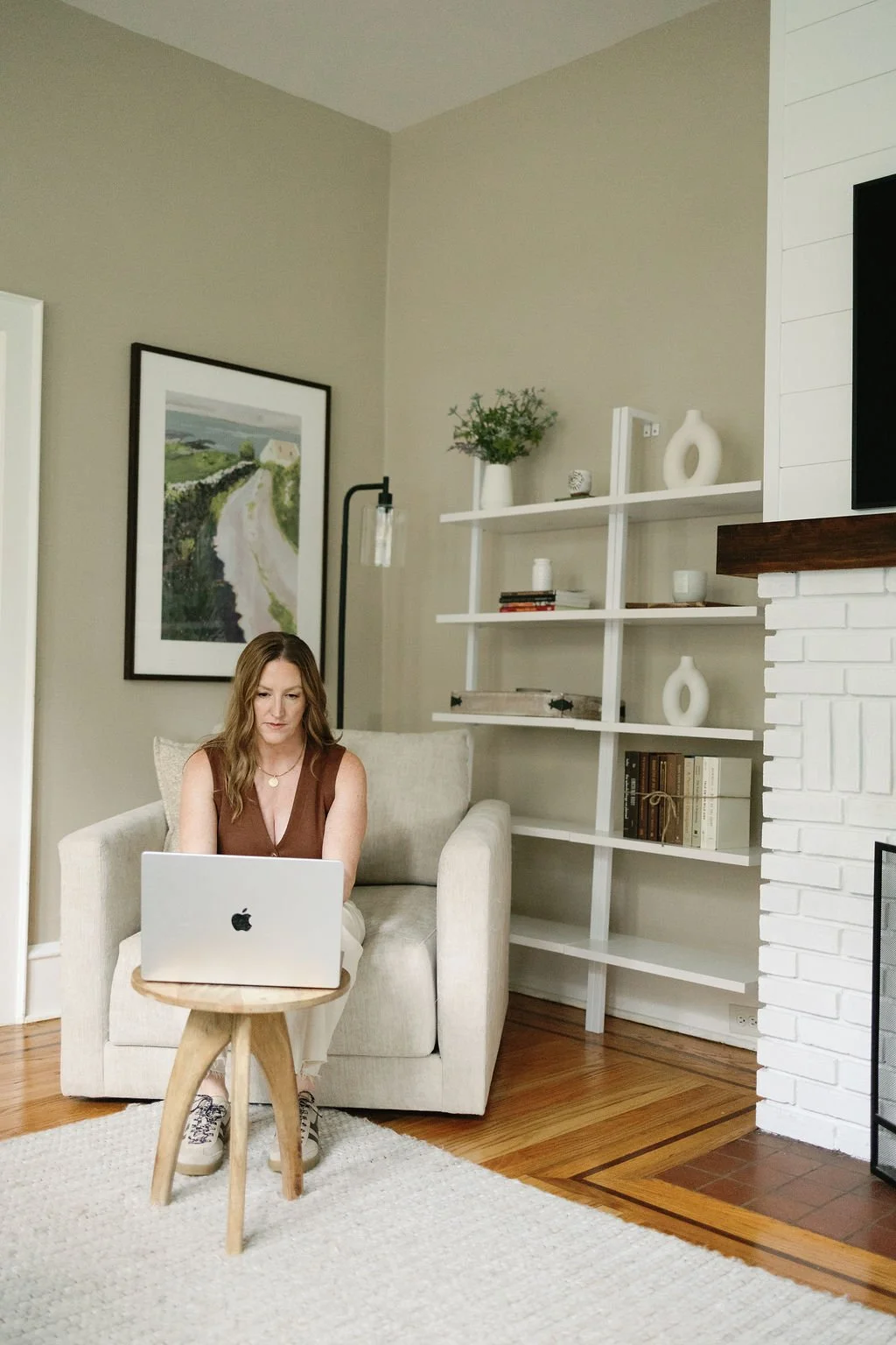 A woman sitting on a beige armchair, working on a silver laptop, in a living room with light-colored walls, a white bookshelf with decorative items and books, a framed landscape painting, a white brick fireplace, hardwood flooring, and a small wooden side table.