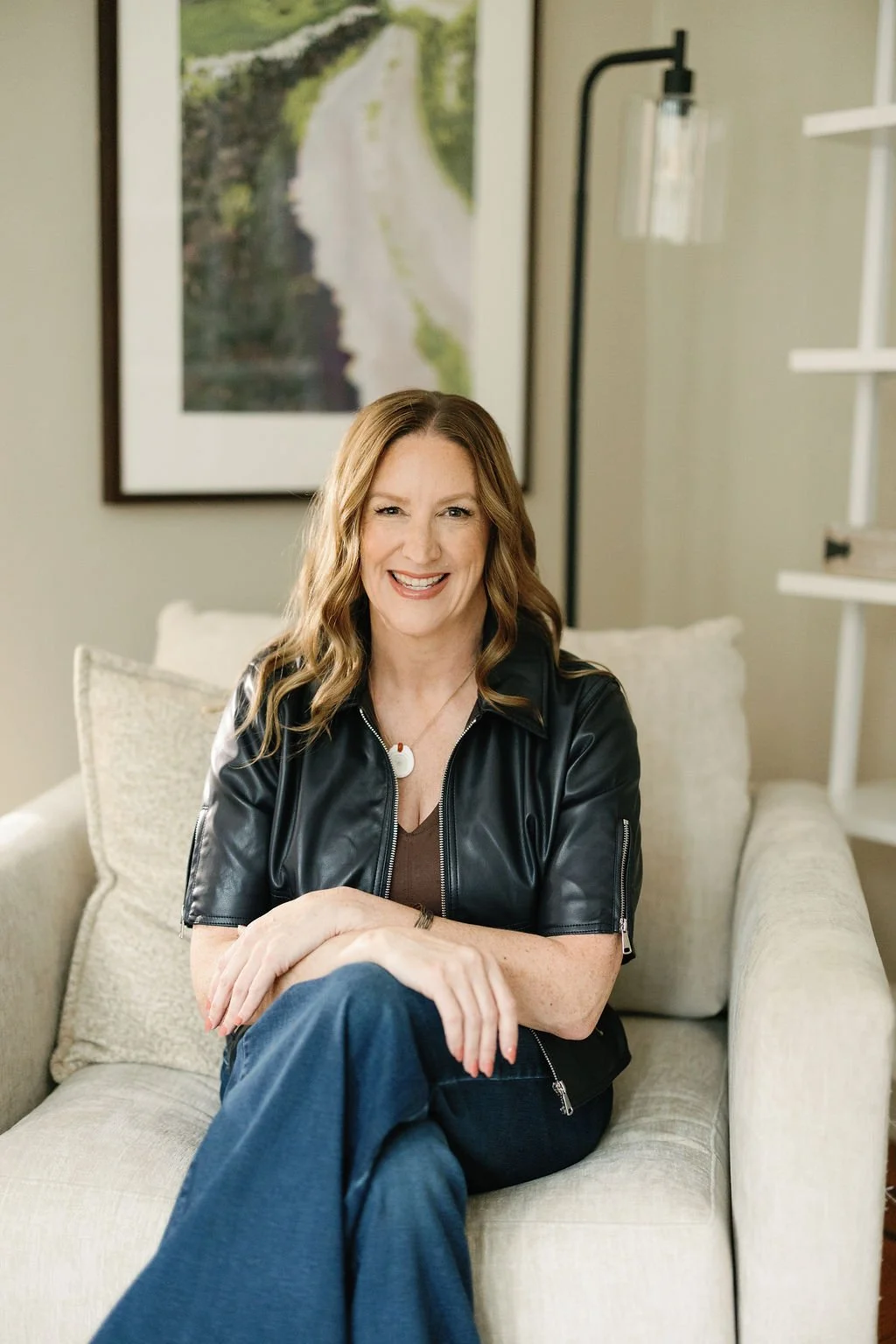A woman with shoulder-length wavy hair, smiling, sitting on a beige couch with crossed legs, wearing a black leather jacket and blue jeans, in a living room with a framed picture of a river and greenery on the wall behind her.