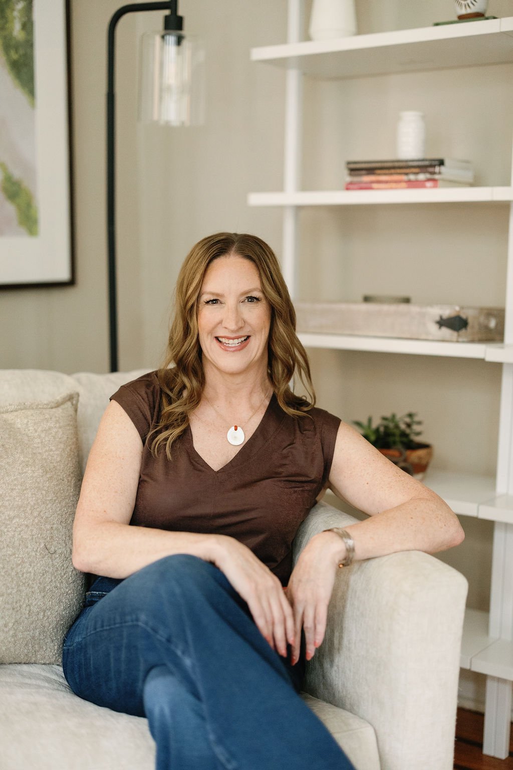 A woman with long, wavy brown hair sitting on a beige sofa, smiling at the camera. She is wearing a brown sleeveless top, blue jeans, a silver watch, and a white pendant necklace. There are white shelves with books and decorative items behind her in a well-lit room.