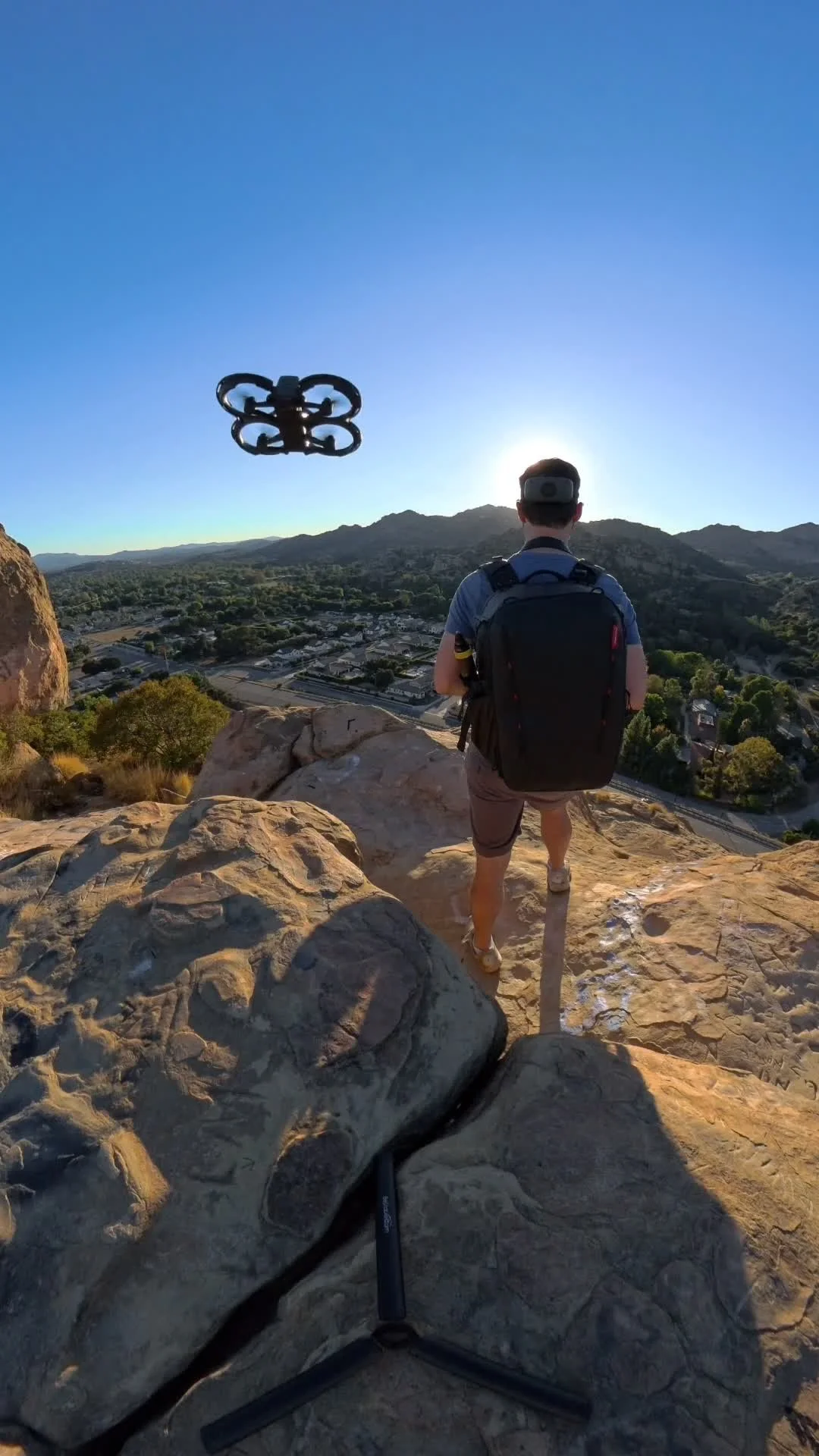 Person standing on rocky ledge overlooking town and mountains, with drone flying nearby at sunset.