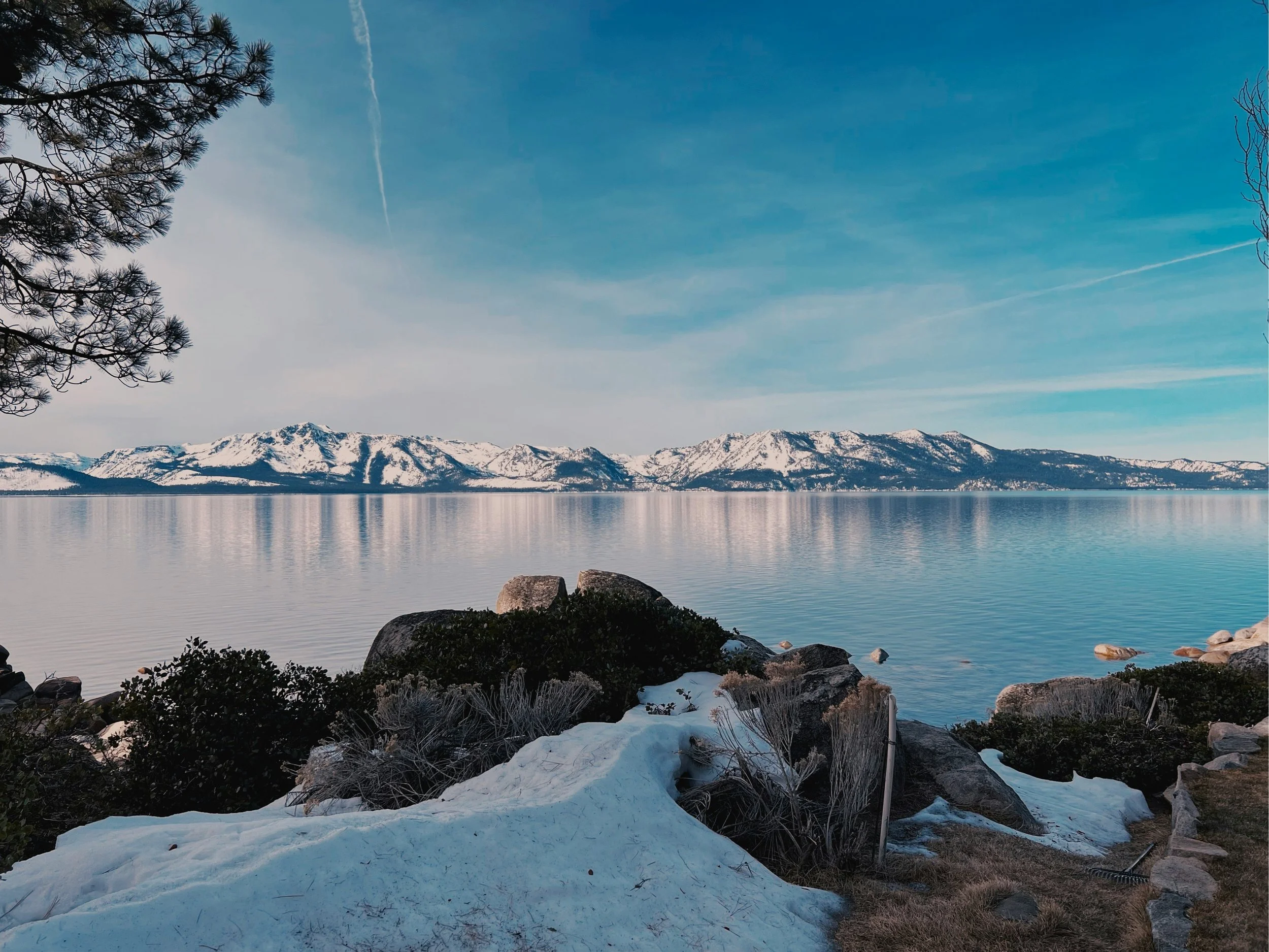 A peaceful lakeside scene with snow covering the ground and rocks in the foreground, pine trees on the left, and a mountain range in the background under a clear blue sky with wispy clouds.