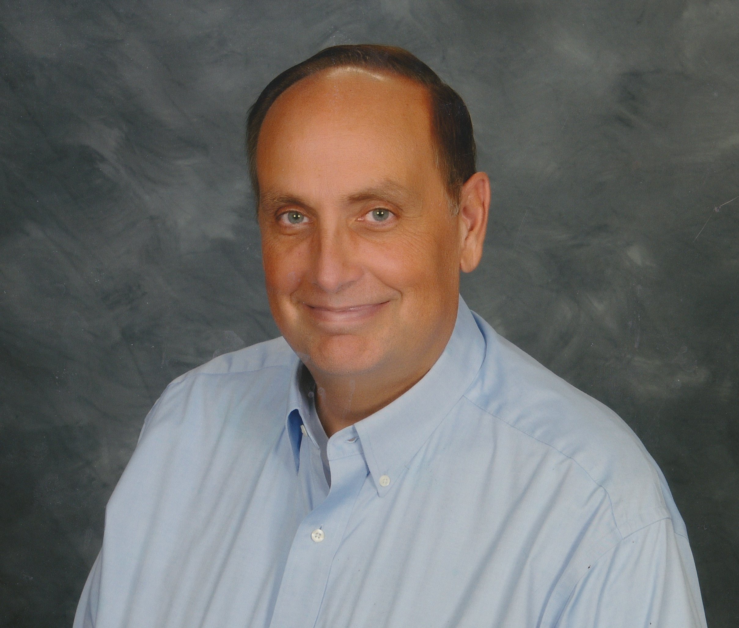 Close-up of a smiling man with light skin, light-colored hair, and blue eyes, wearing a light blue dress shirt, against a dark, textured background.