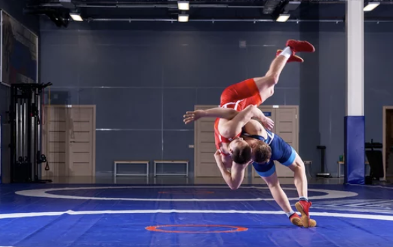 Two female wrestlers in red and blue uniforms wrestling on a blue mat in a gym.