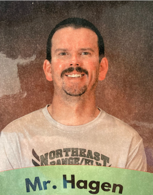 Photograph of a man with dark hair, a mustache, and a beard, smiling. He is wearing a beige T-shirt with black and green text that reads, 'Northeastern' and 'Mr. Hagen' at the bottom. The background is a reddish-brown gradient.