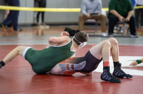 Two young boys wrestling on a red mat in a gymnasium, with spectators sitting in the background.