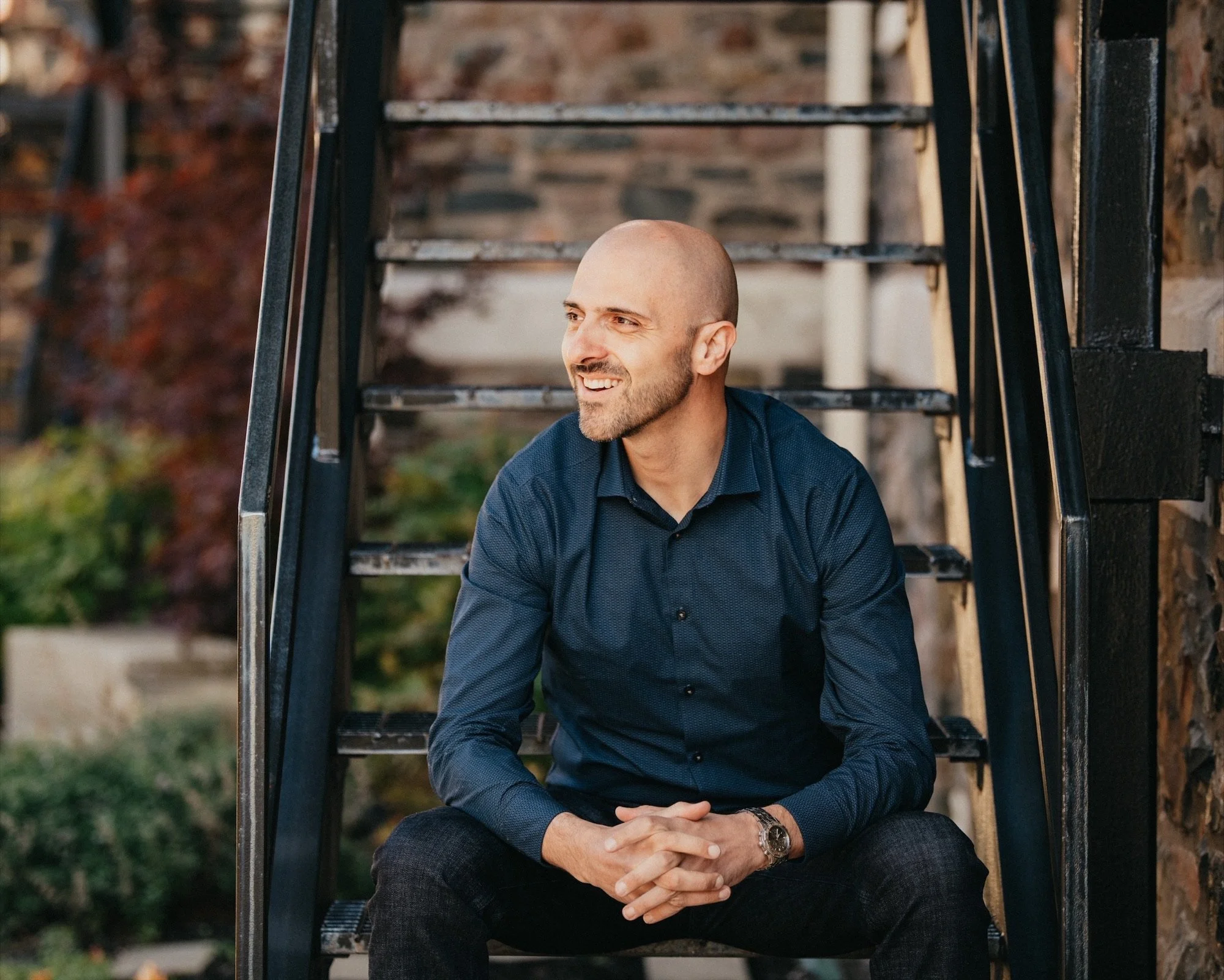 A man with no hair and a beard, wearing a dark blue shirt, sitting on outdoor metal stairs with a brick and foliage background, smiling and looking off to the side.