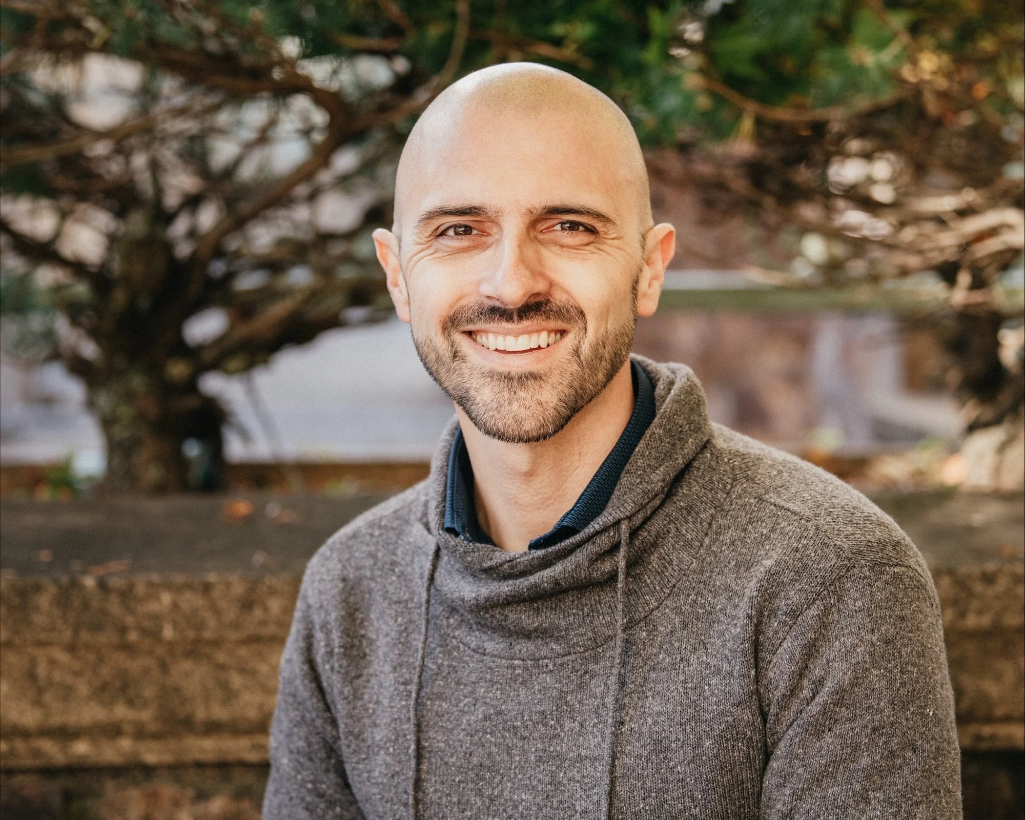 Smiling man with a shaved head and beard outdoors, wearing a gray hoodie, with blurred nature background.