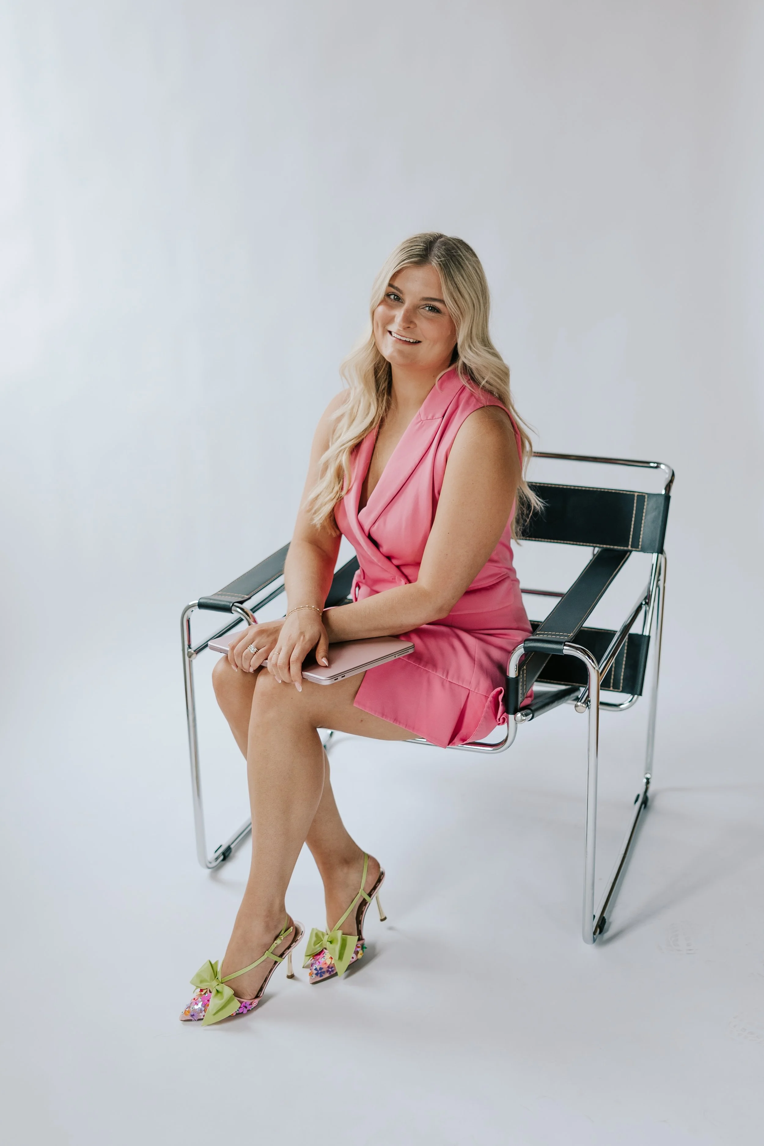 A woman with long blonde hair sitting on a modern black and chrome chair against a plain white background. She is smiling, wearing a pink dress, and colorful high heels with floral and bow details.