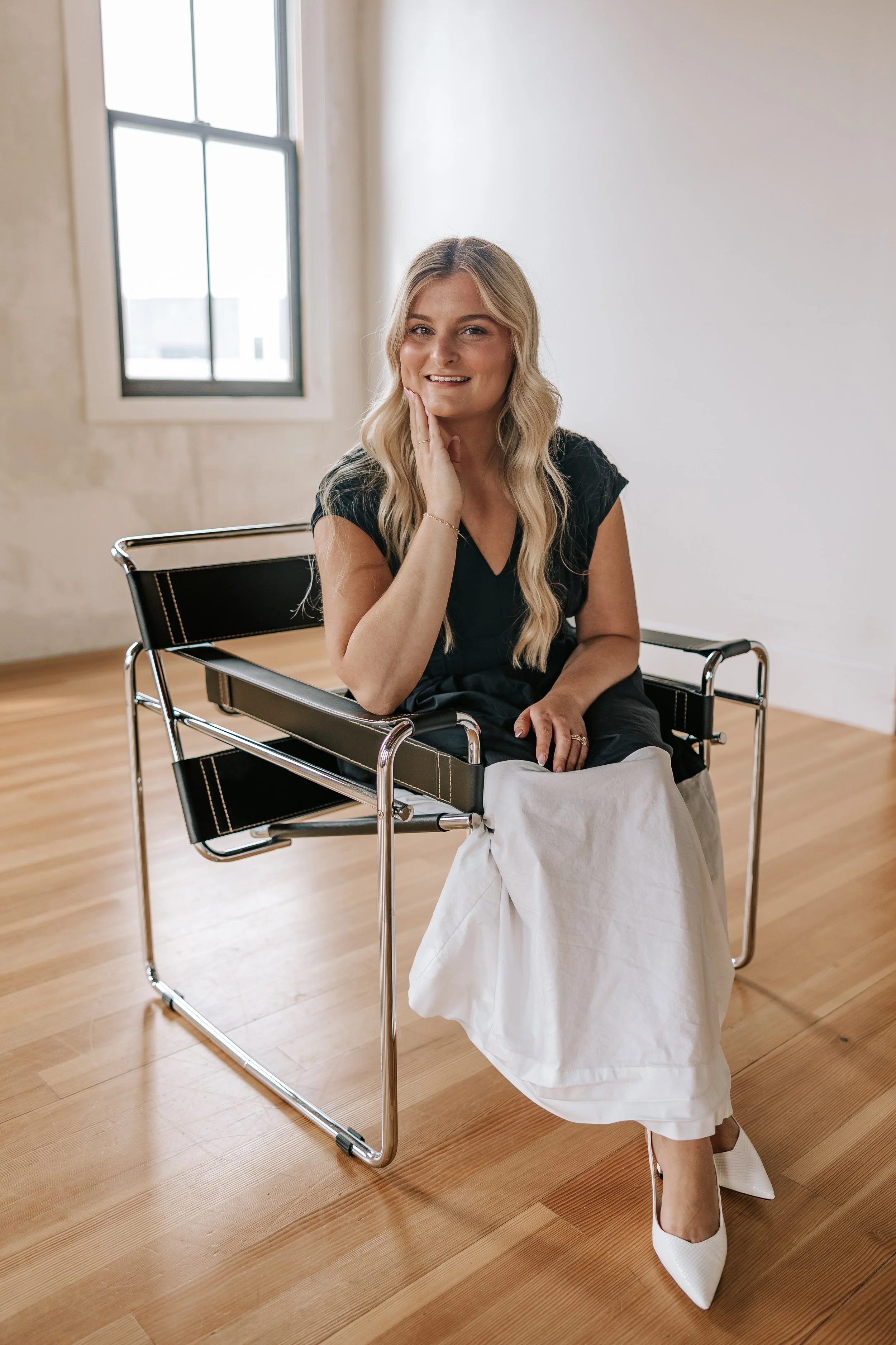 A woman with long blonde hair smiling while sitting in a black and chrome chair in a room with wooden floors and large windows.