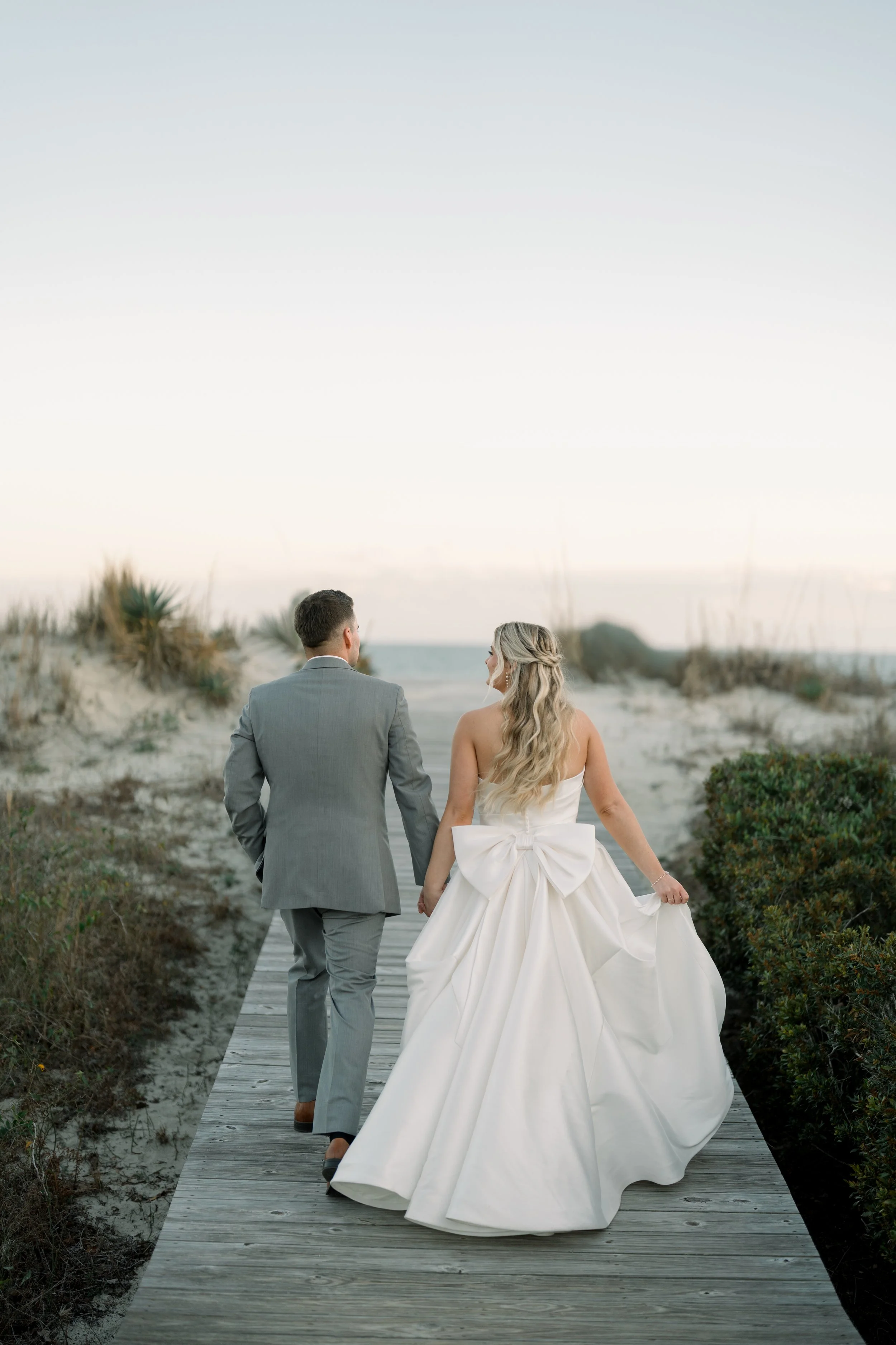 A bride and groom walking hand in hand on a wooden boardwalk through a beach landscape at dusk.