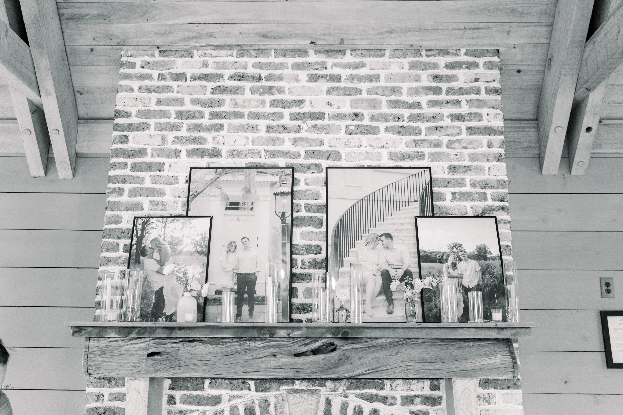 A display of multiple framed black and white photographs of a couple, arranged on a wooden shelf against a brick wall and wooden ceiling beams.