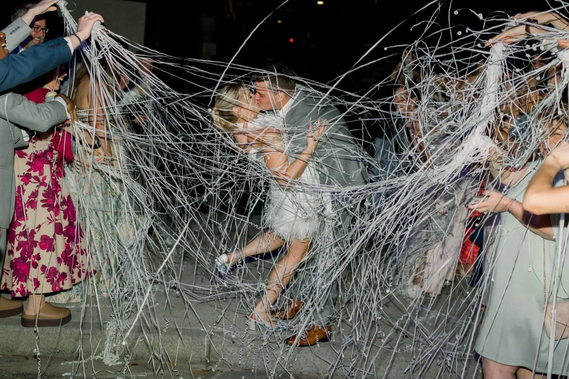 A bride and groom kissing amid guests at their wedding reception, surrounded by streamers and decorations.