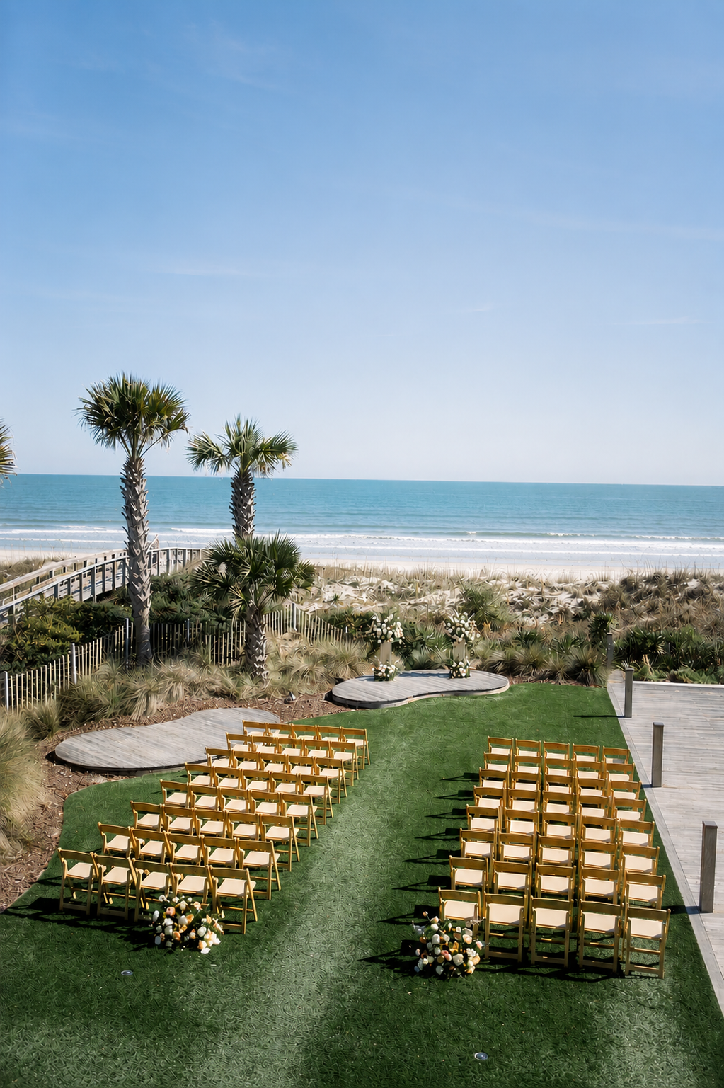 Beachside wedding ceremony setup with yellow chairs arranged on a green grassy aisle, decorated with flower arrangements, overlooking the ocean with palm trees in the background on a sunny day.