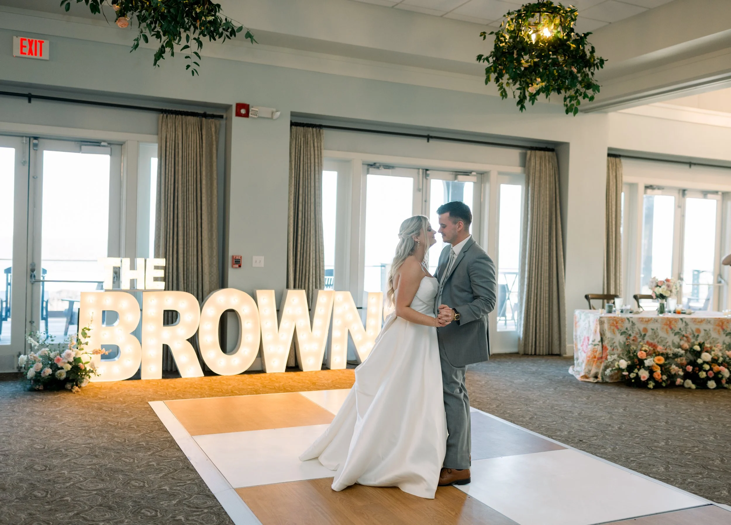 A bride and groom dancing at their wedding reception indoors. Large illuminated letters spell out 'THE PROM' in the background. The room has large windows, curtains, and floral decorations on a table to the right.