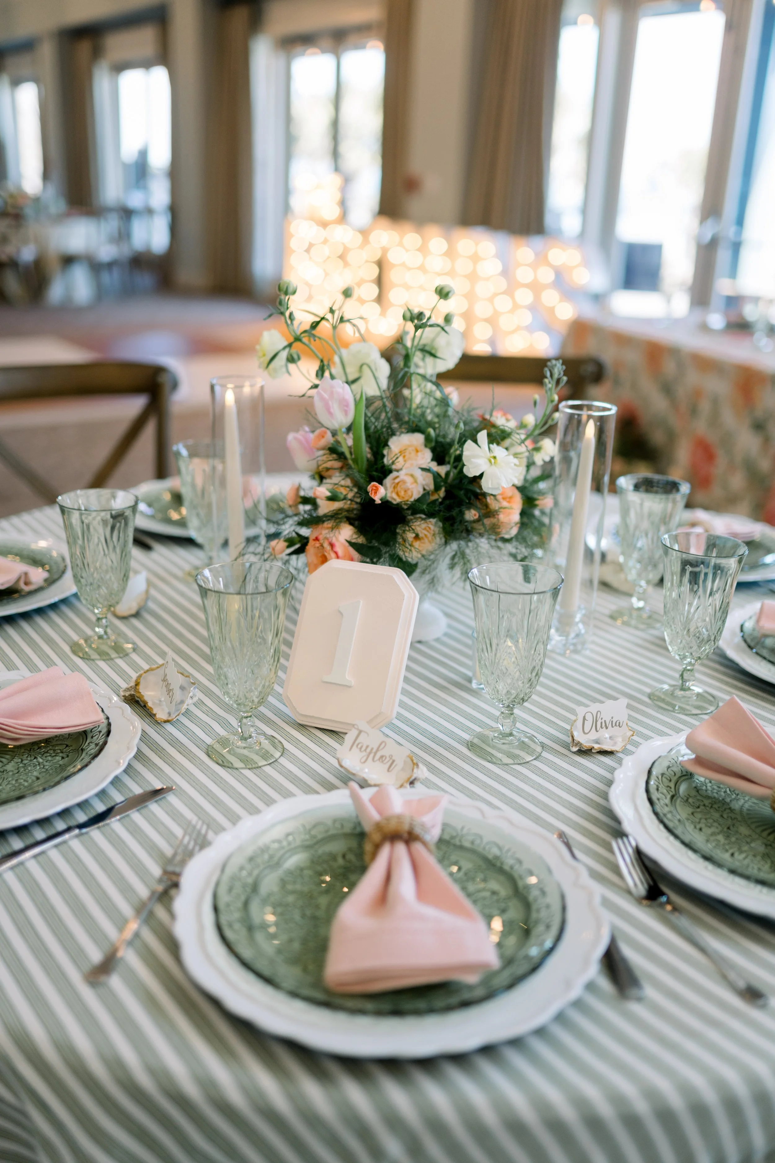 A decorated dining table with a floral centerpiece, glassware, plates with green patterned layers, pink napkins folded into a bow, and name tags, set for a celebration or event in a well-lit room with large windows and string lights in the background.
