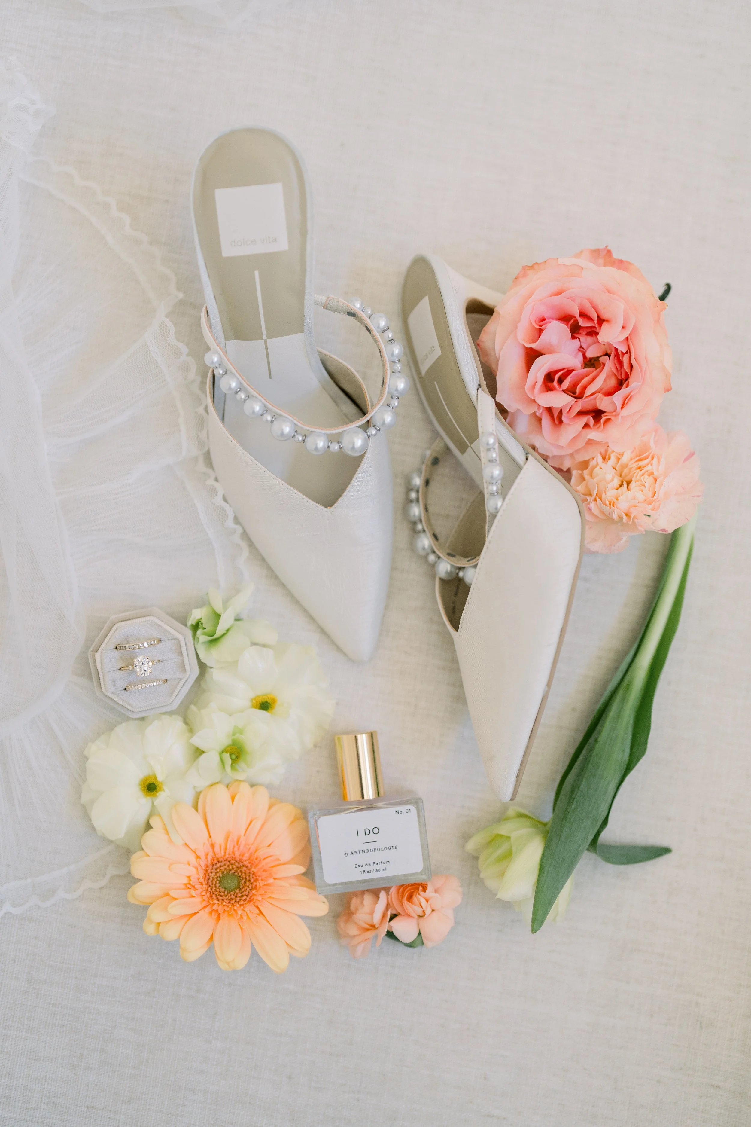 A display of white high-heeled shoes decorated with pearls, a pink flower, and various flowers and perfume bottles on a white fabric surface.