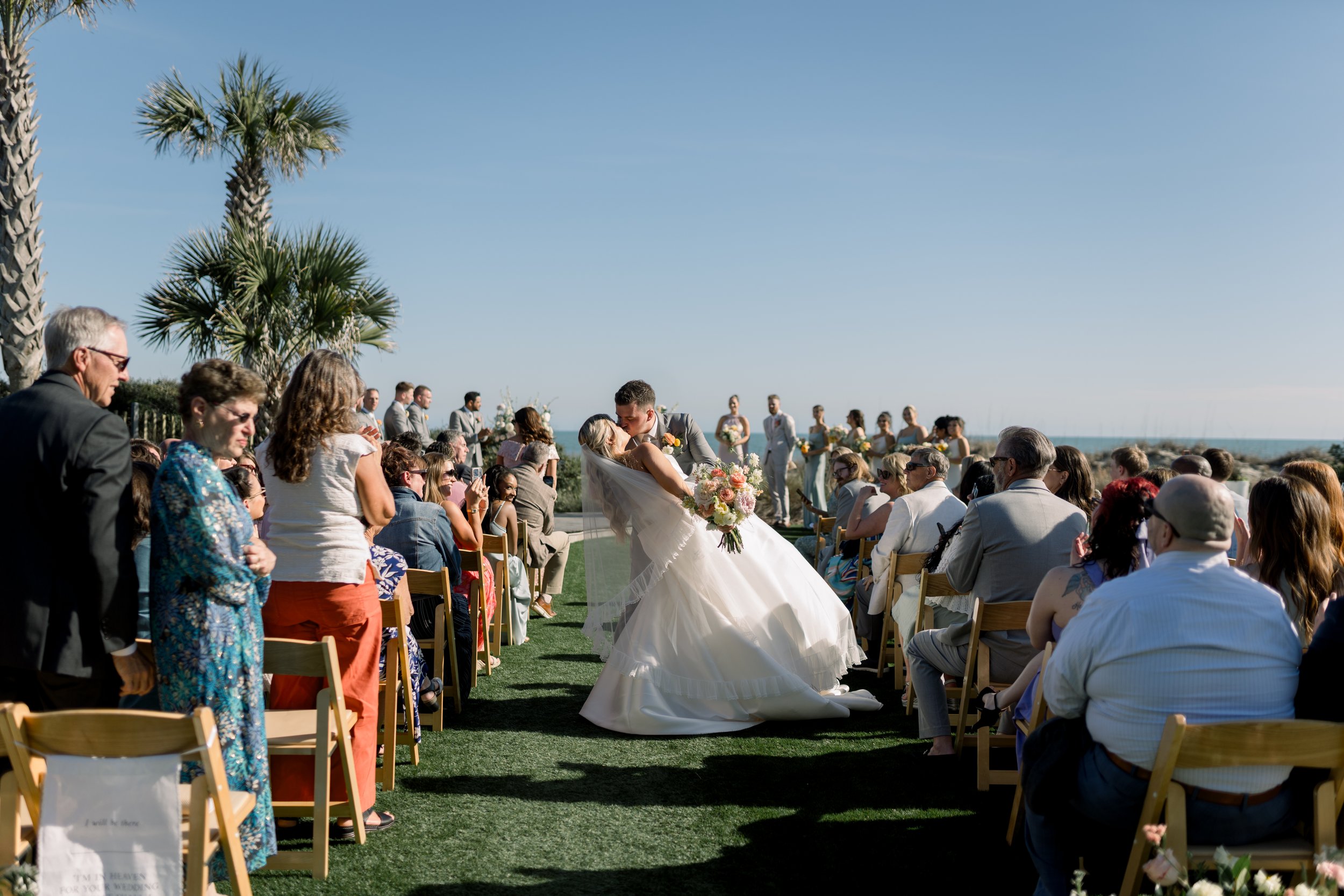A wedding ceremony on a beach. The bride and groom are kissing, surrounded by guests seated on chairs. The bride is holding a bouquet of flowers, and the ocean is visible in the background under a clear blue sky. Palm trees are also present.