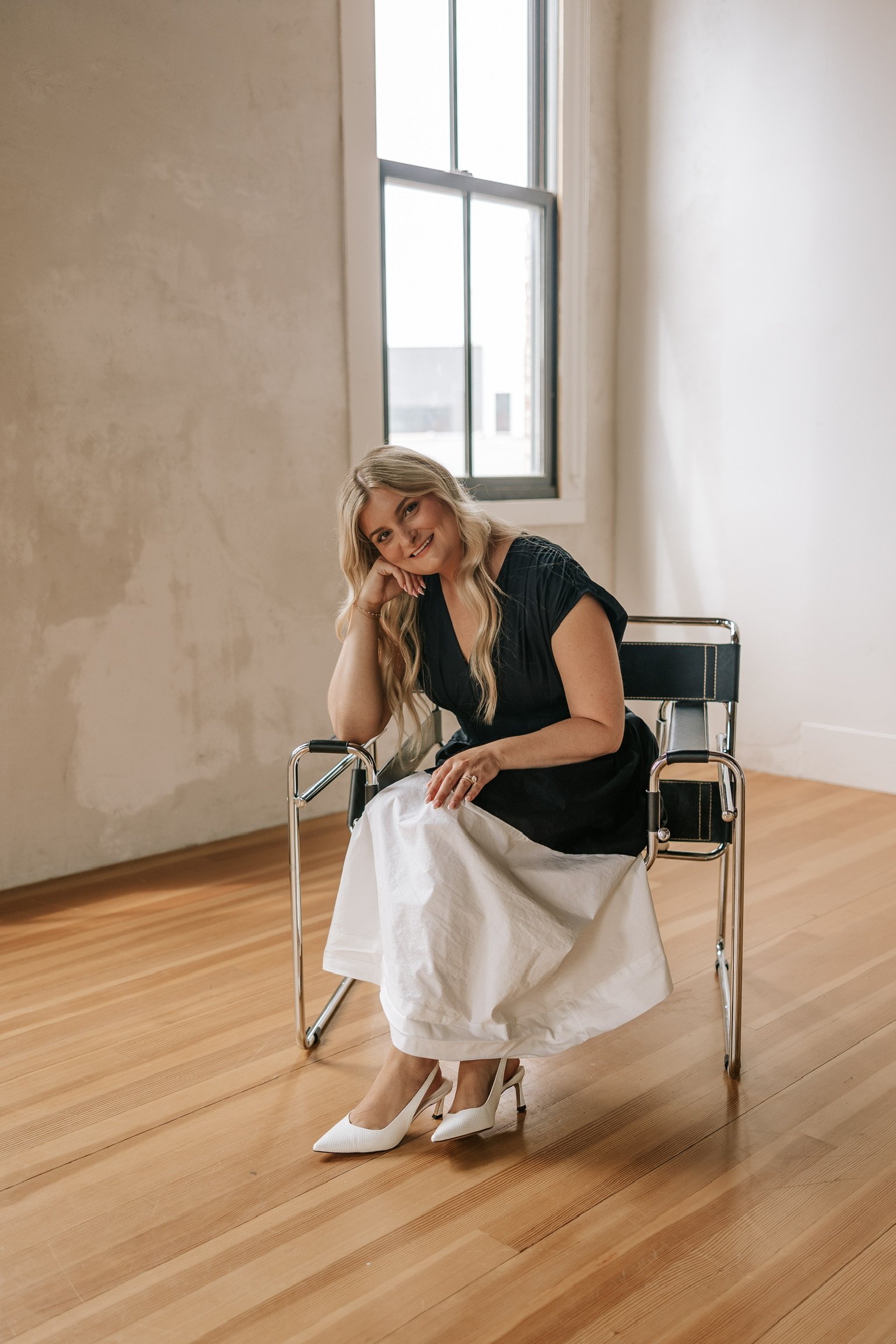 A woman with blonde hair sitting in a wheelchair in a bright room with wooden floors and large windows. She is smiling, resting her head on her right hand, wearing a black top, white skirt, and white high heels.