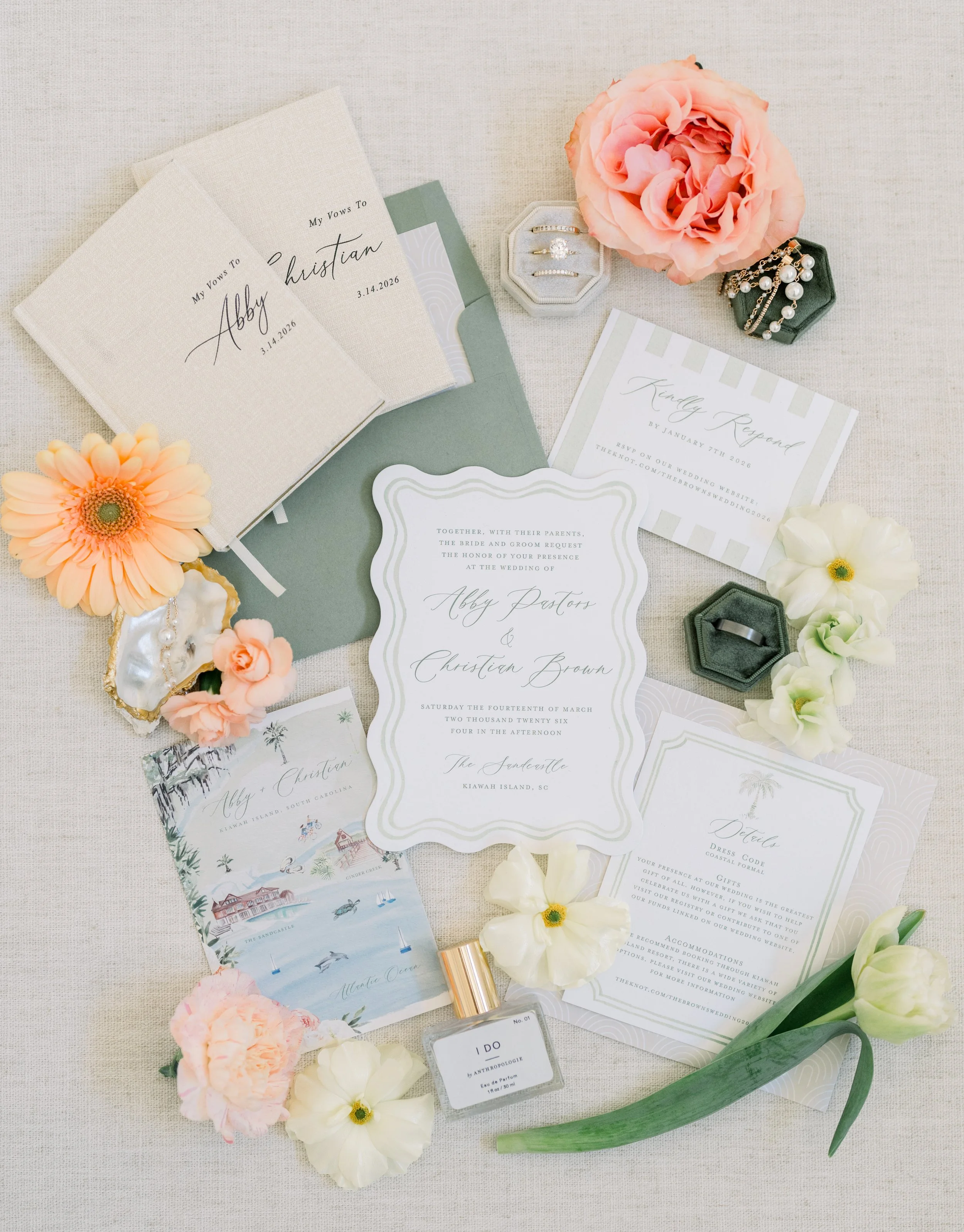 Wedding invitation suite with floral decorations, including a pink rose, white and blush flowers, pearl jewelry, rings, perfume, and travel brochures, arranged on a white fabric background.