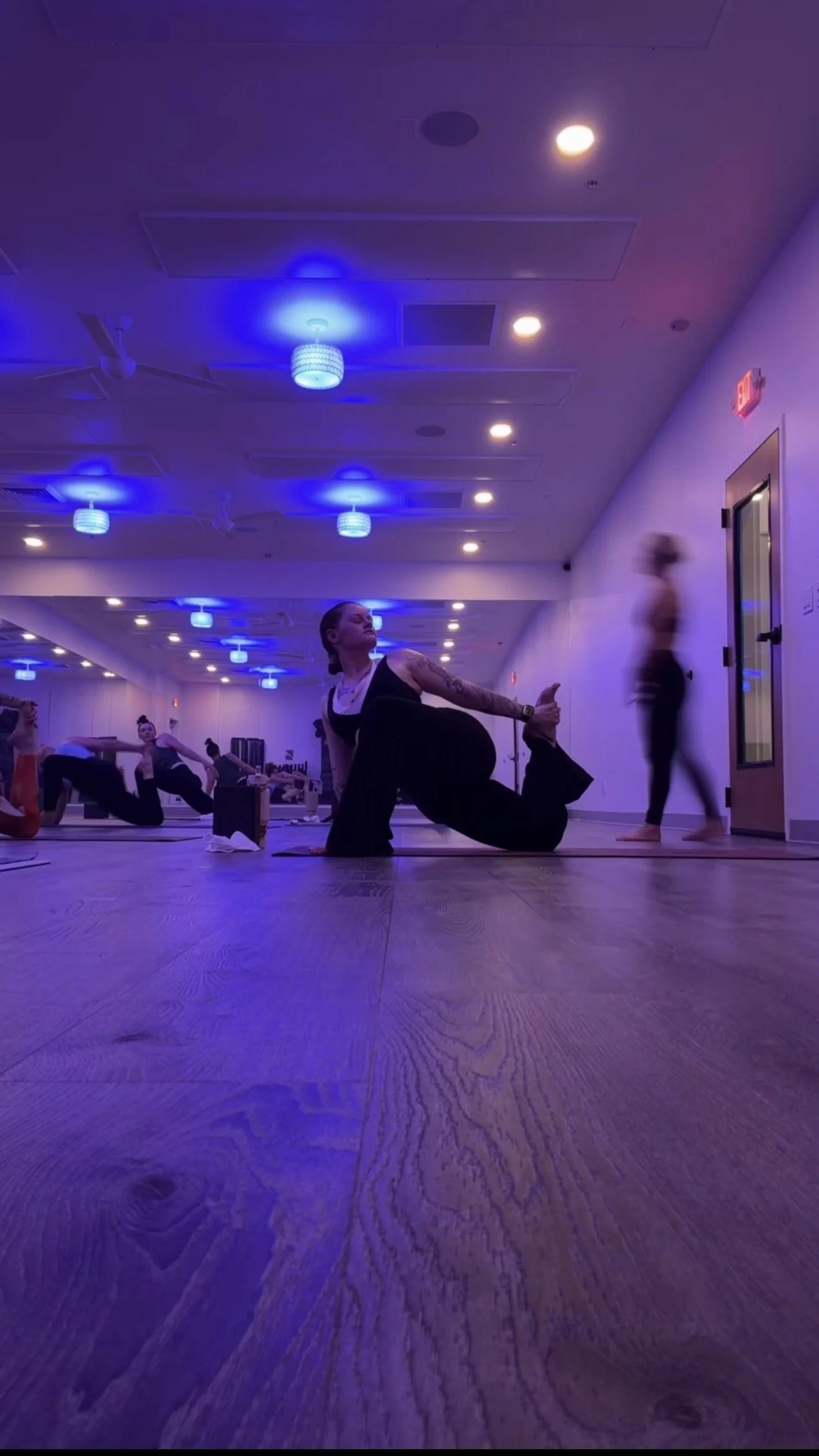 A woman practicing yoga in a dimly lit studio with blue lighting and mirrors, surrounded by other participants, and a person walking by in the background.