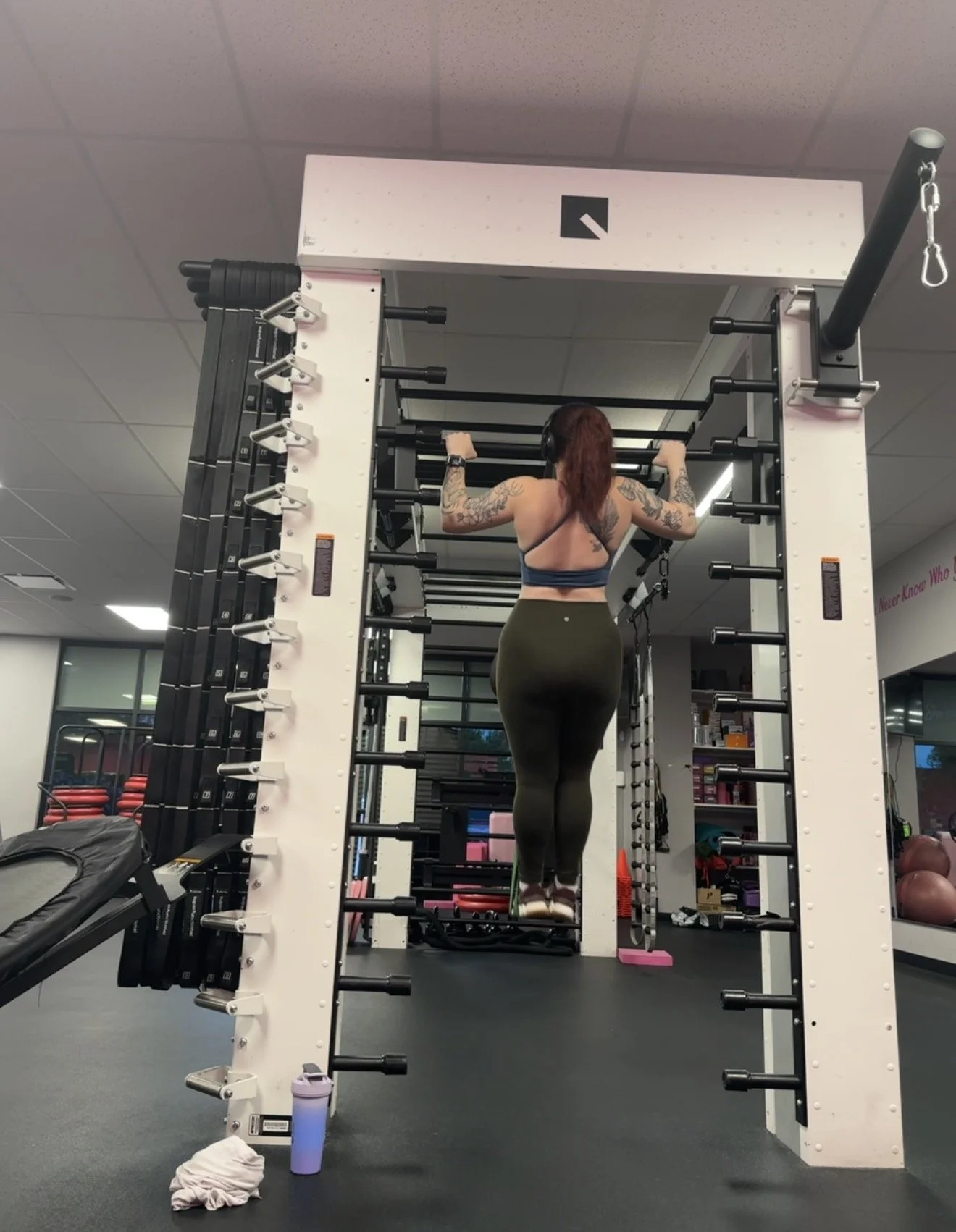 A woman with tattoos is doing pull-ups on a gym machine in a gym. She is wearing a sports bra and leggings.