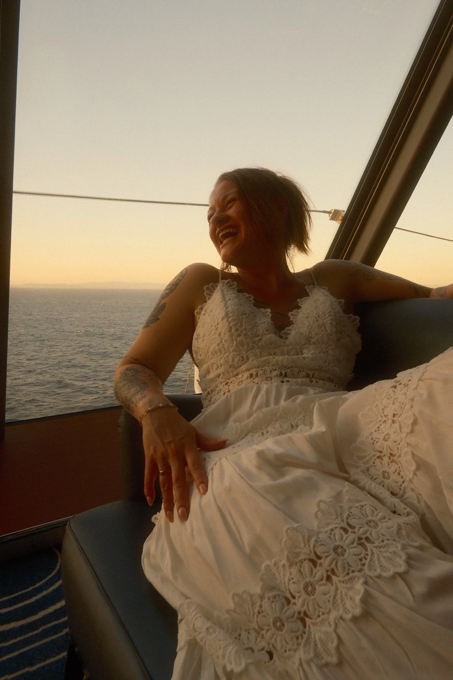 A woman with tattoos on her arm sitting by a window, smiling and laughing, wearing a white lace dress, with a view of the ocean at sunset in the background.