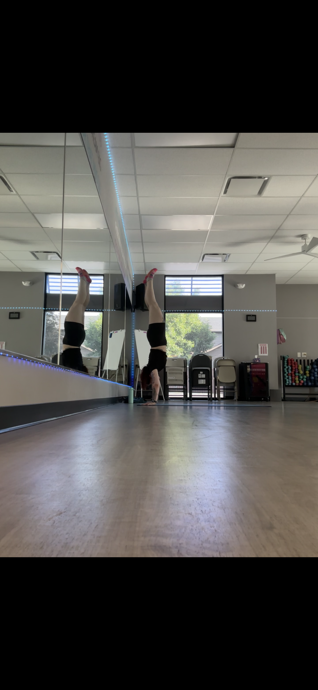 A woman performing a handstand in a dance or fitness studio, with her reflection visible in a large mirror on the wall.