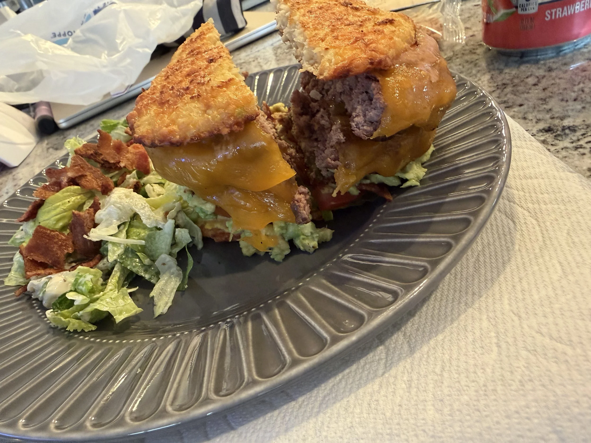 Half of a cooked cheeseburger with lettuce and tomato on a metal plate, with some crumbled bacon and a side salad, on a granite countertop.