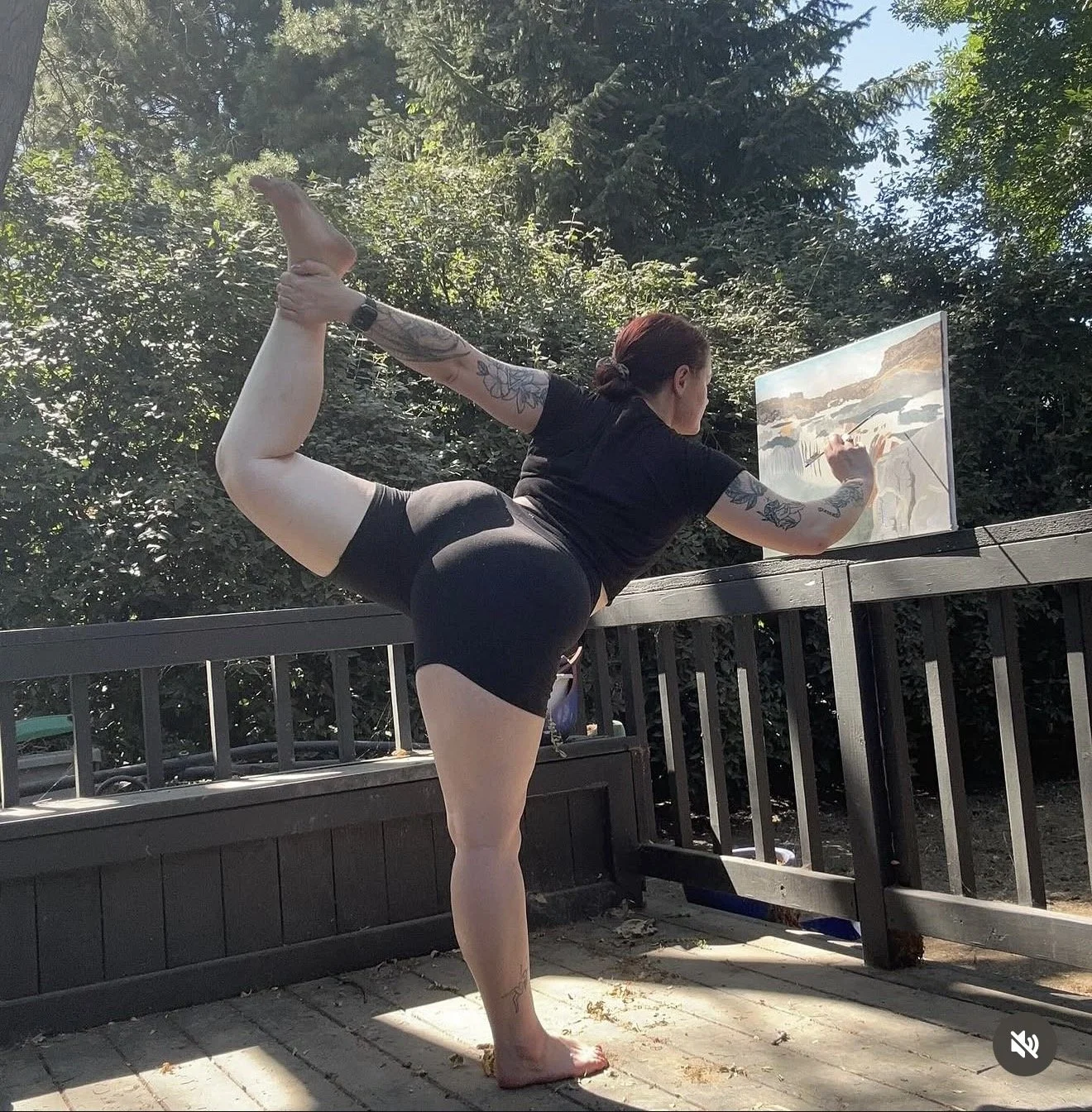 A woman with tattoos doing a yoga pose on a wooden deck outdoors, balancing on one foot while holding her other foot behind her back with one hand and painting on a canvas with the other hand.