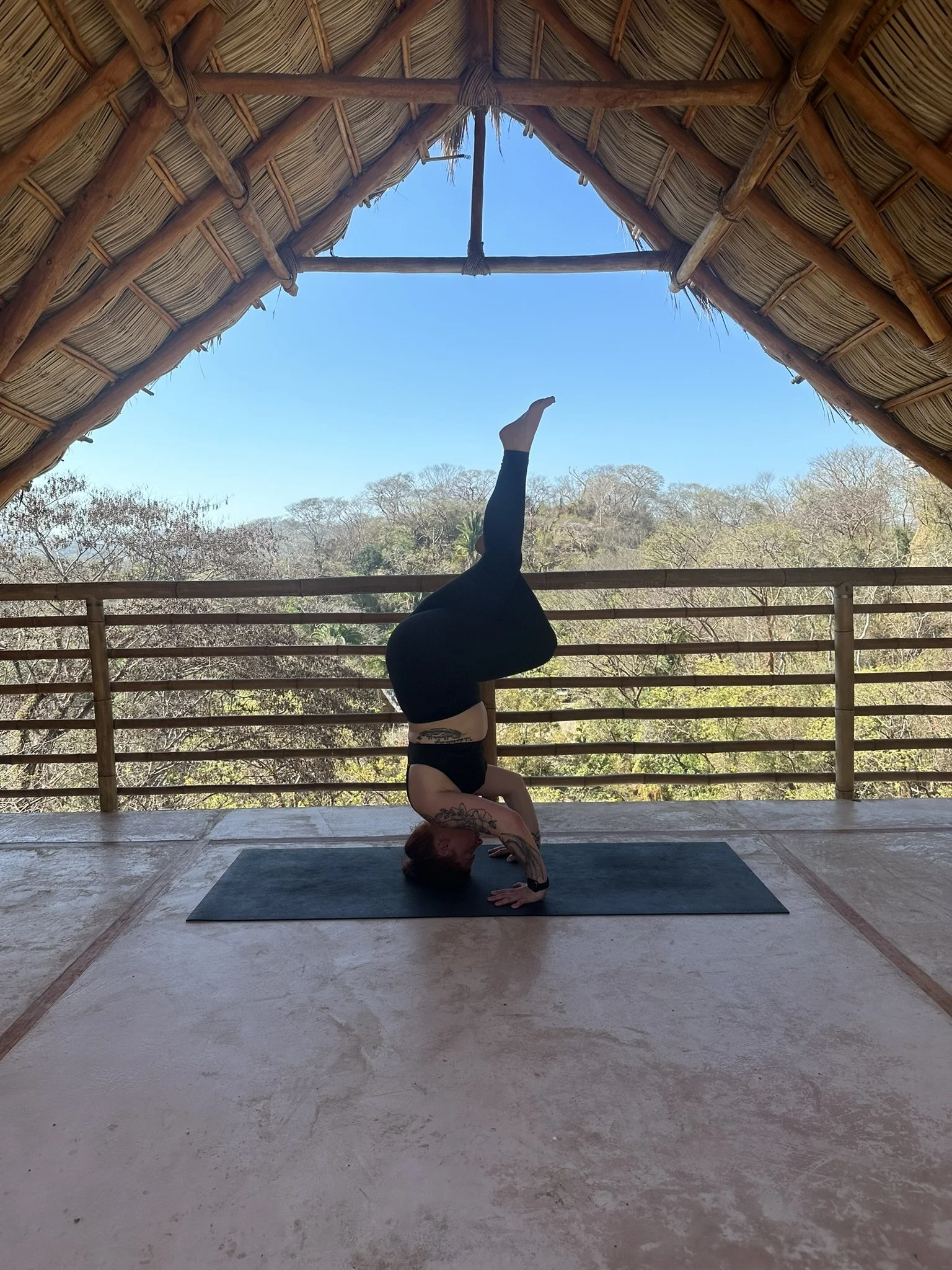 A person practicing yoga in a headstand pose on a black mat inside a bamboo structure with a thatched roof, overlooking trees and a clear blue sky.