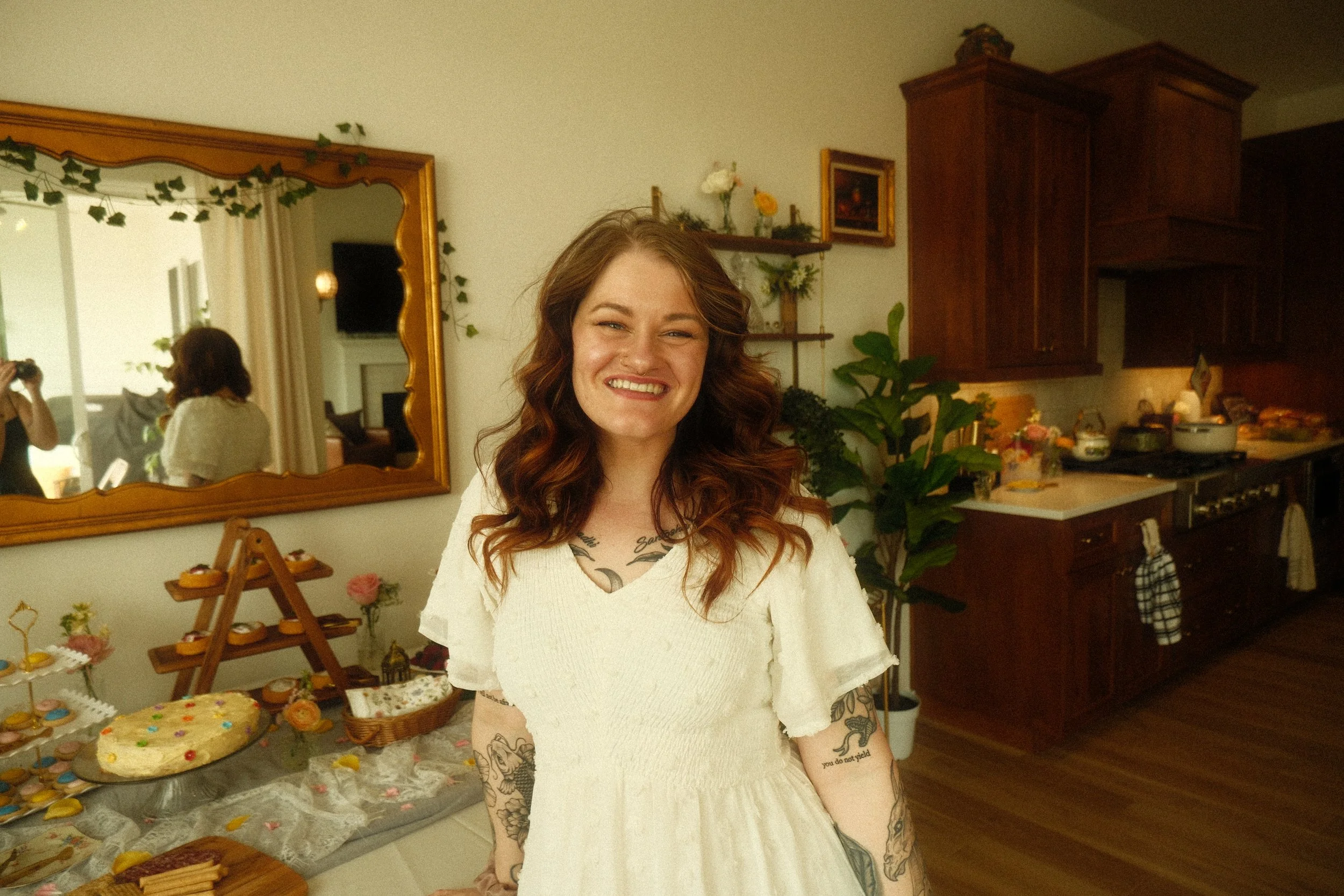 A woman with tattoos and wavy hair smiling in a kitchen decorated for a celebration, with a table of cakes and cookies in the background.