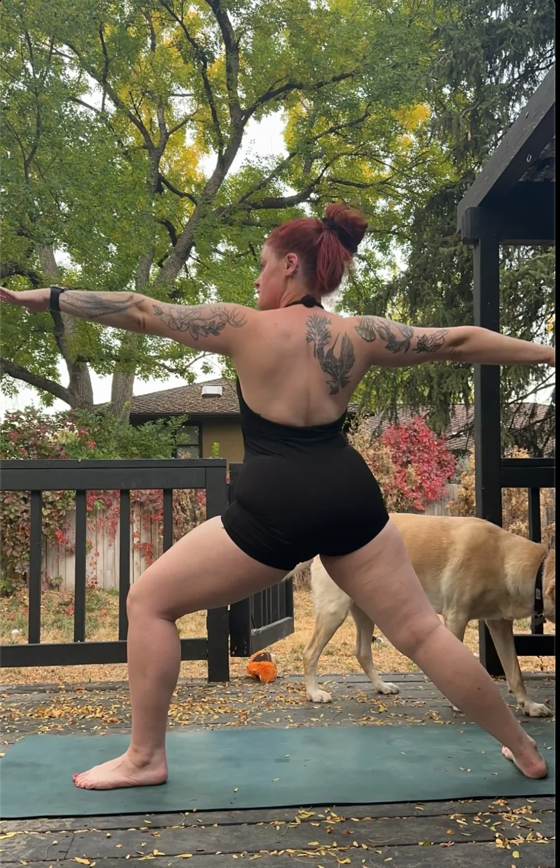 A woman with tattoos practicing yoga outdoors on a green mat, with a dog walking in the background, in a backyard with trees and a wooden fence.