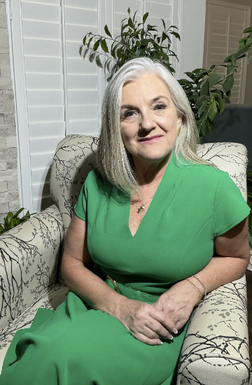 An older woman with gray hair sitting on a patterned armchair in front of a large leafy plant in a room with white shutters.
