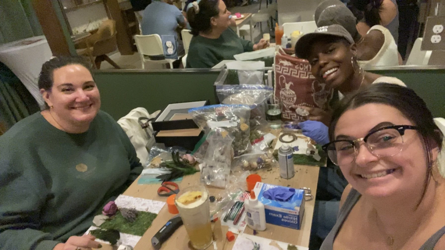 Four women smiling at a table filled with craft supplies, including scissors, glue, and decorations, in a busy indoor setting.