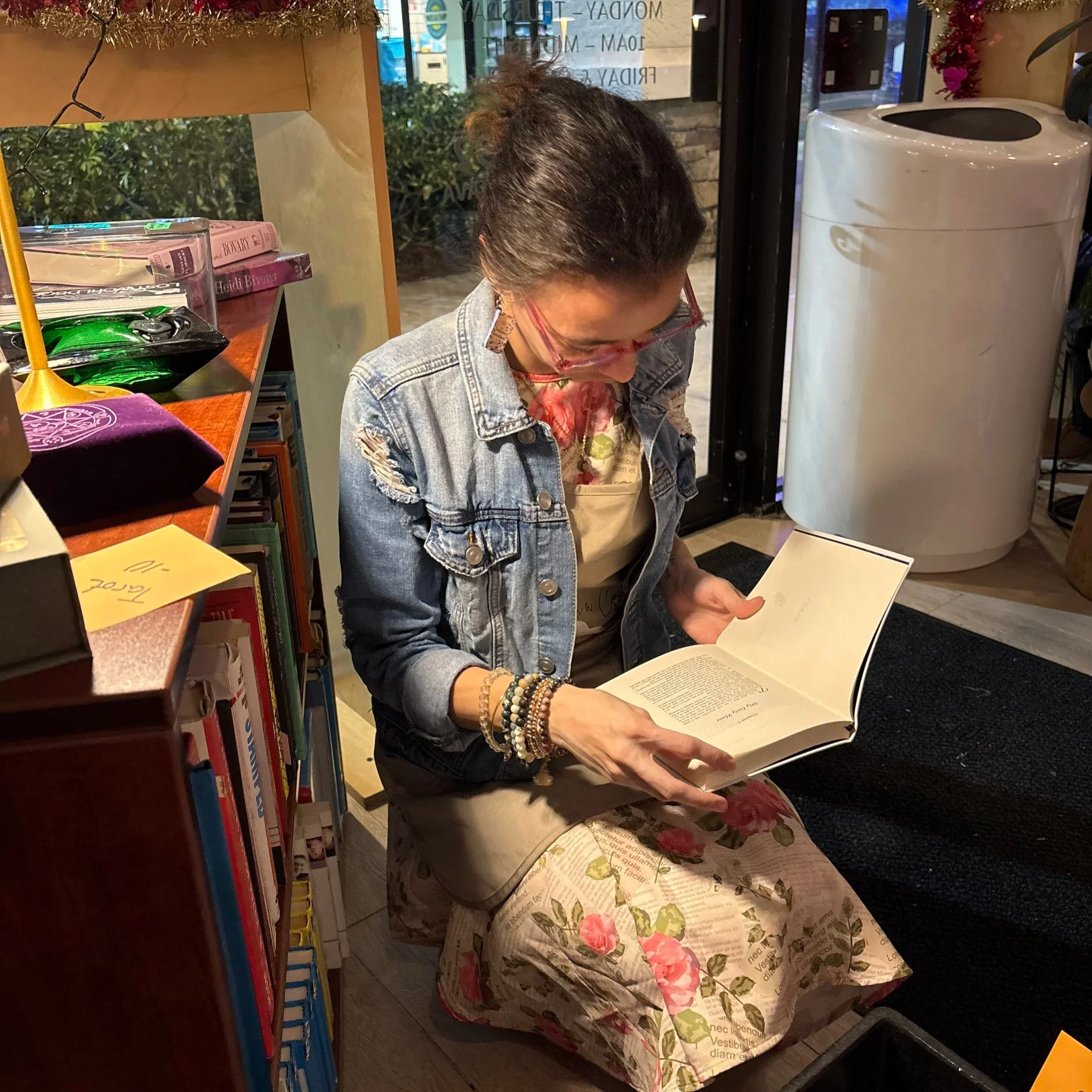 A woman reading a book while sitting on the floor in a bookstore or library, near a bookshelf with books and a counter with a floral tablecloth at a SWFL Creatives hosted event.