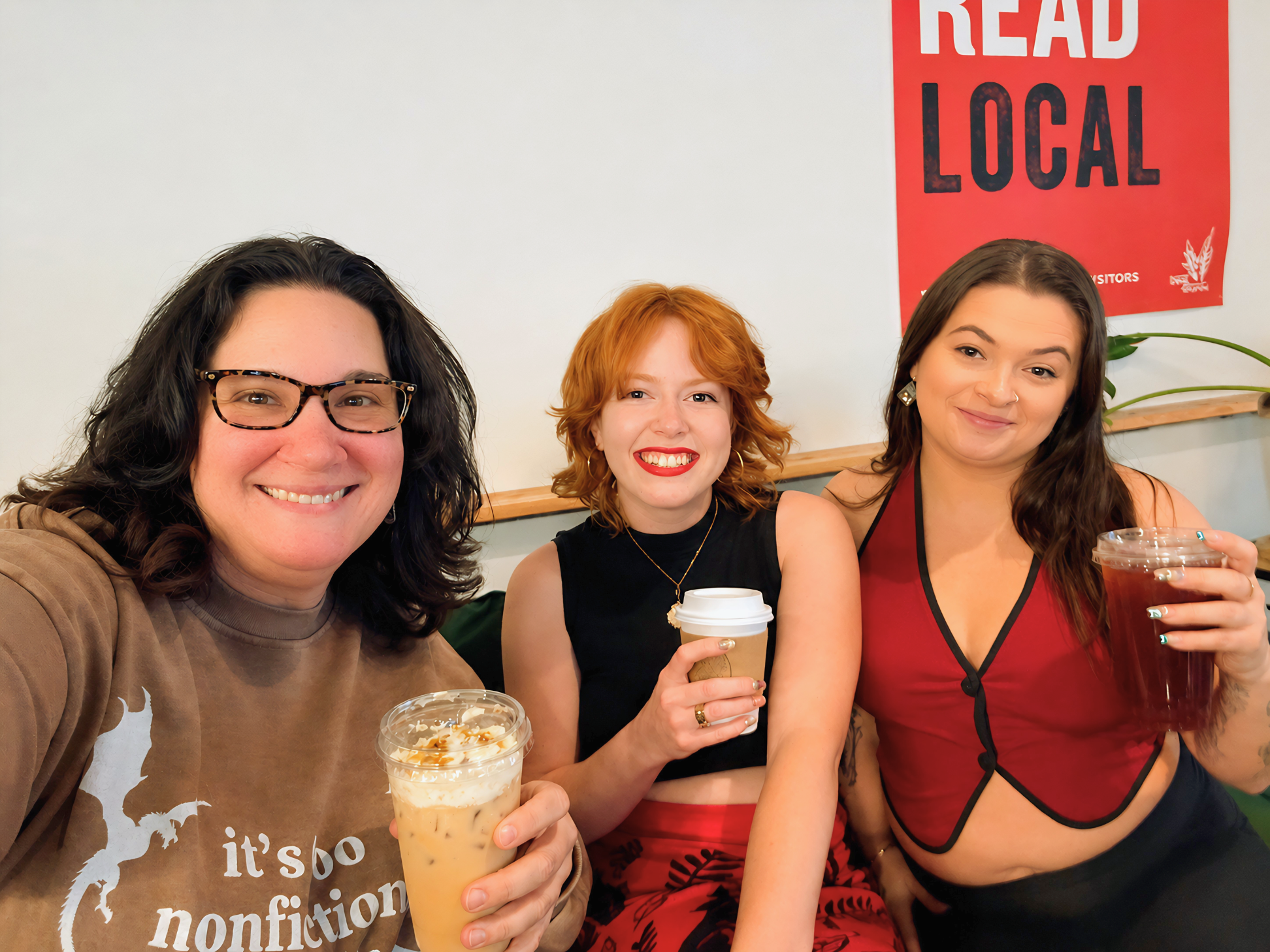 Three founding women of SWFL Writers and Creatives smiling and holding iced coffee drinks, sitting together at a social gathering. A red poster with white and black text reading 'READ LOCAL' is visible in the background.