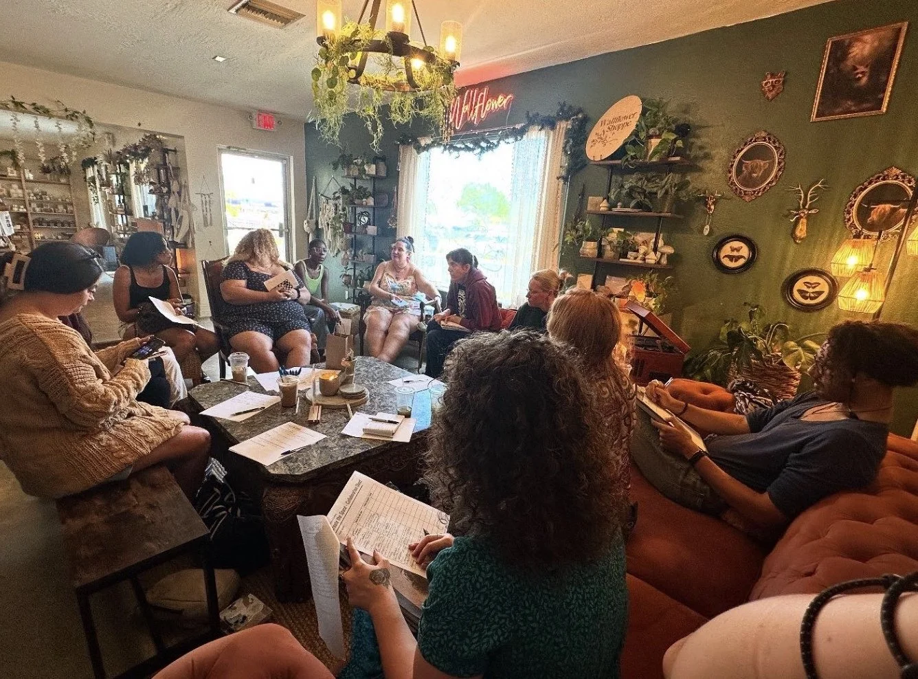 A group of women sitting in a cozy, decorated room with plants, artwork, and warm lighting, engaged in a meeting about community events.