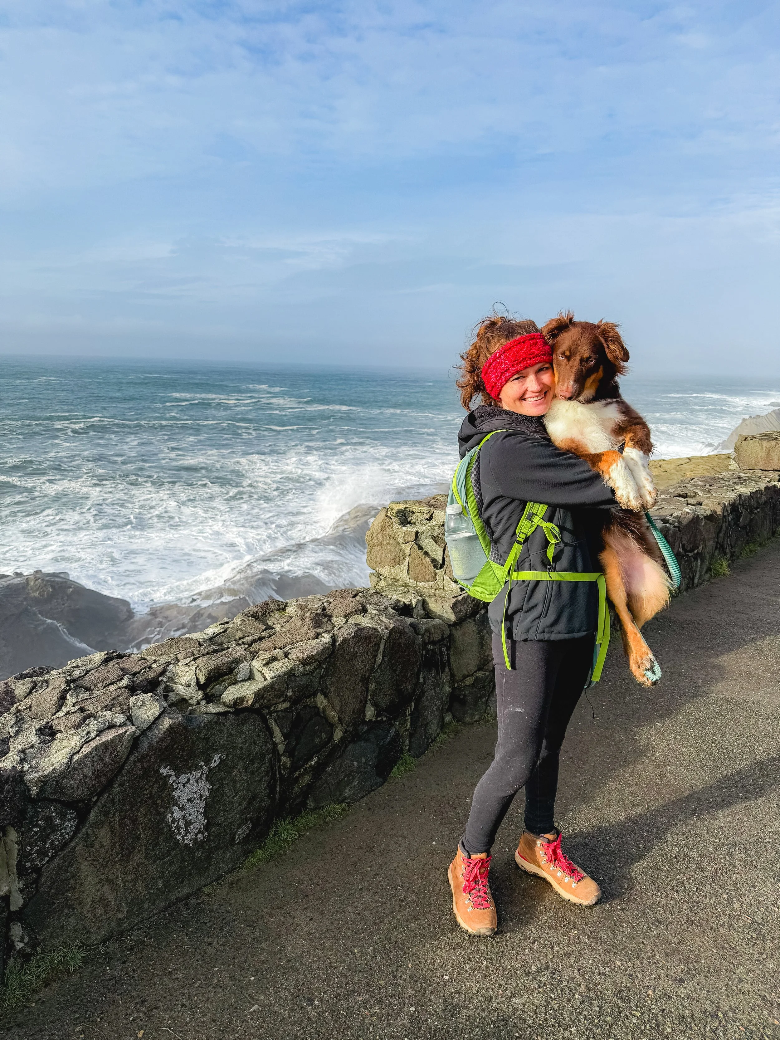 A woman in hiking gear holding a dog by the coast with an ocean and sky in the background.