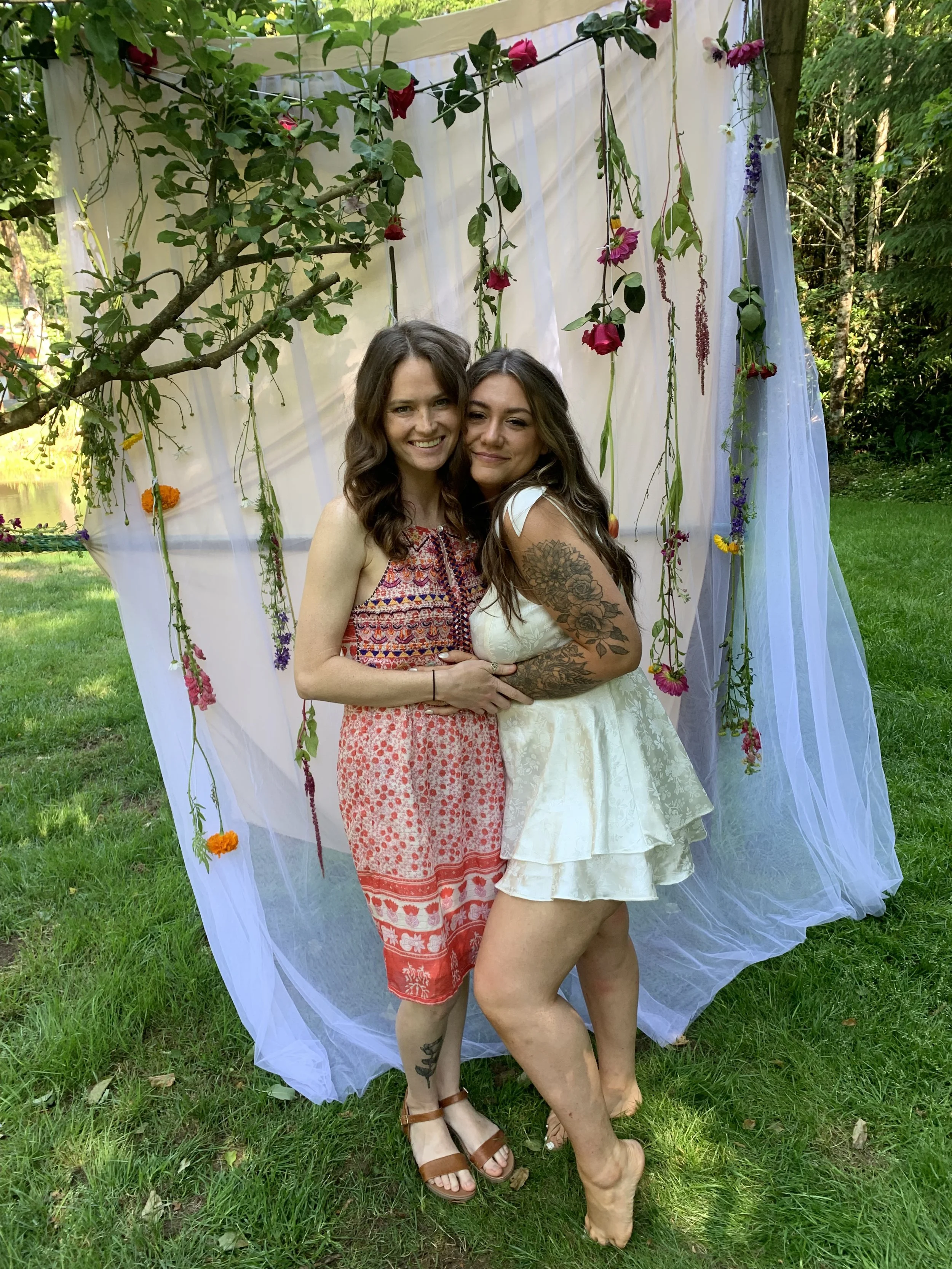 Two women are standing close together outdoors in front of a decorative backdrop with hanging flowers and sheer white fabric. They are smiling and embracing each other.